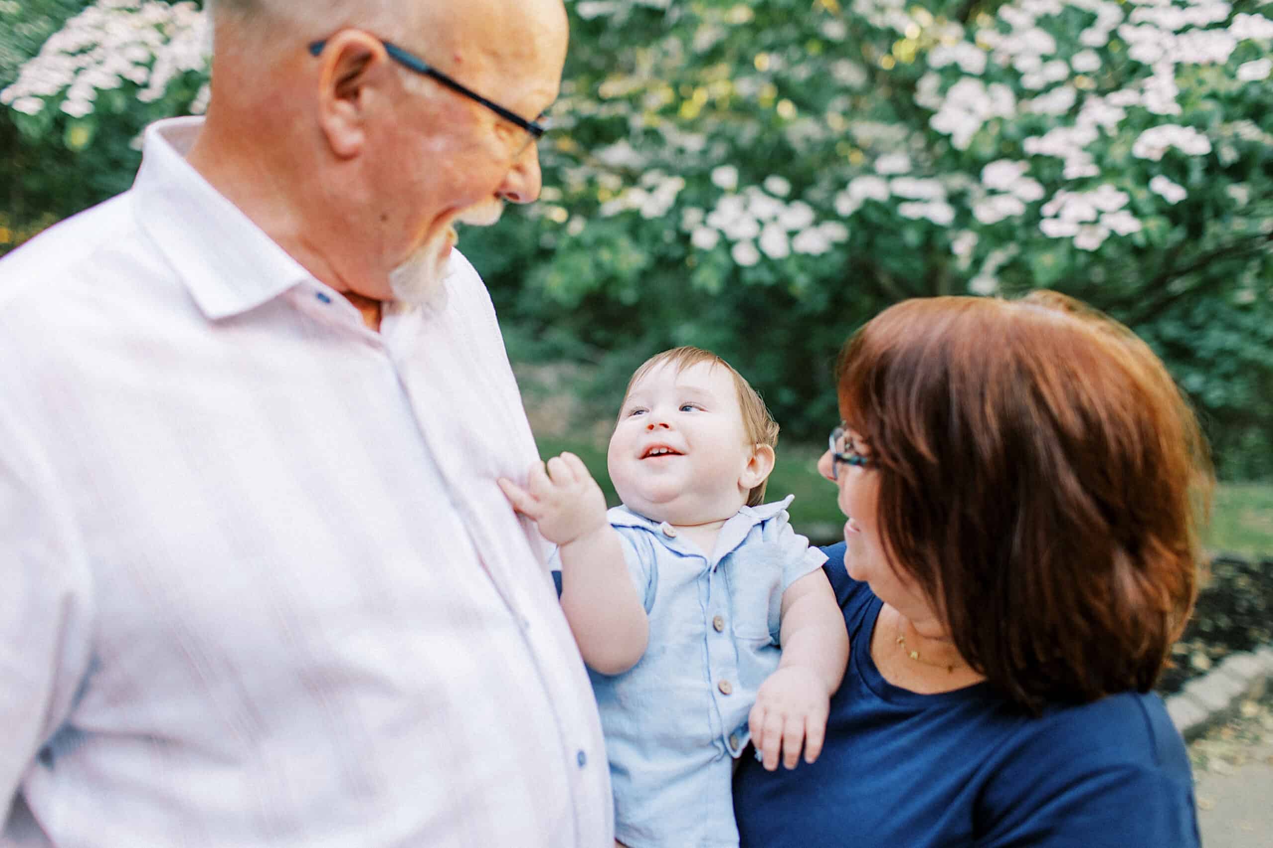 A man and woman hold a smiling baby outdoors, surrounded by green foliage and flowering plants—perfect for Spring Family Photos in Bucks County.