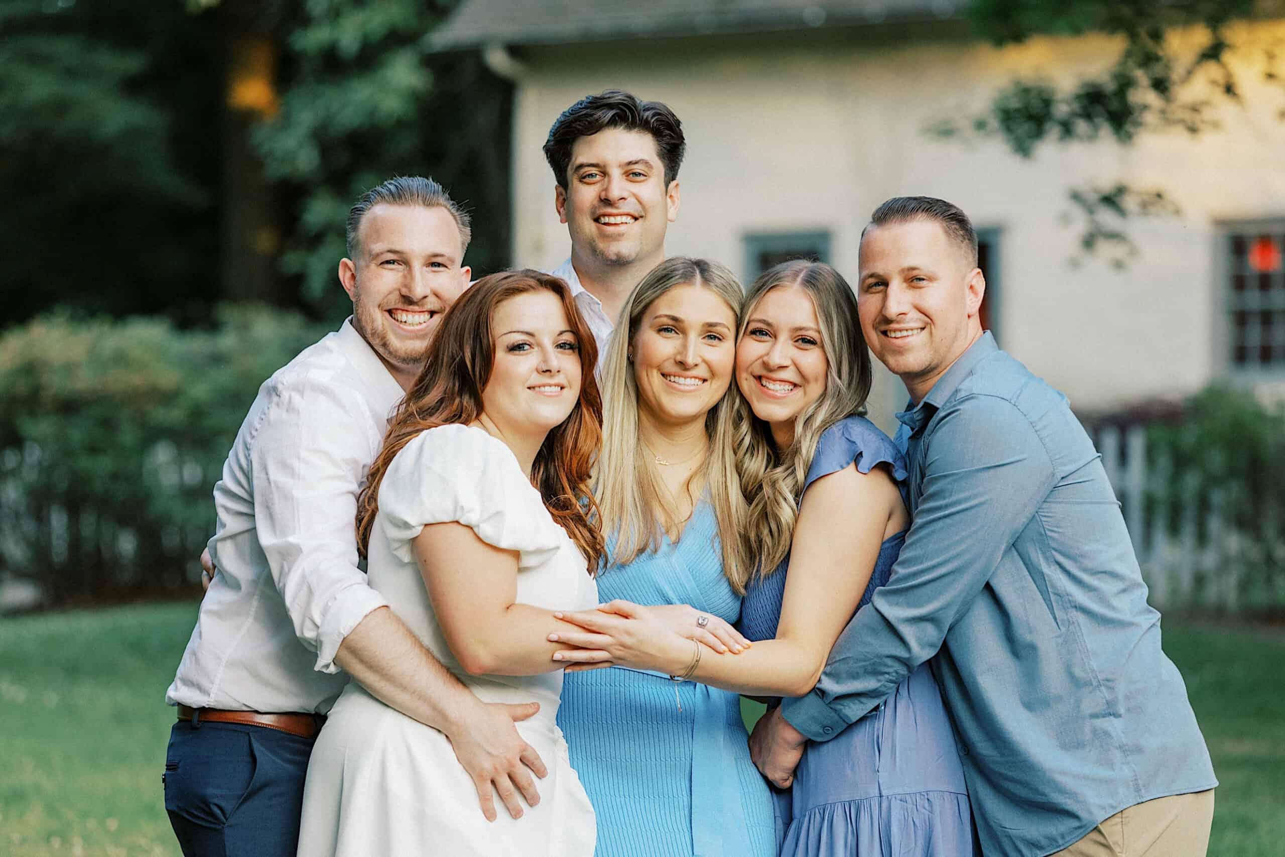 Six adults stand closely together outdoors, smiling at the camera, with greenery and a house in the background—capturing the warmth of Spring Family Photos in Bucks County.