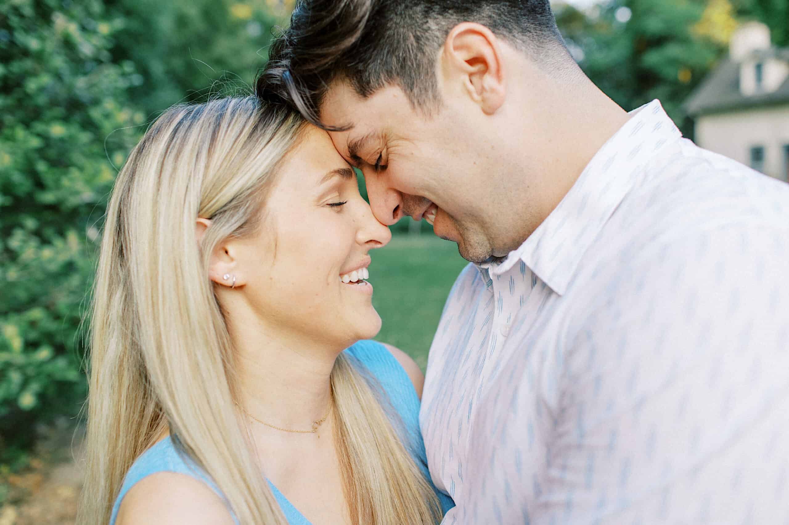 A man and woman stand close together outdoors, touching foreheads and smiling, with lush greenery and a house in the background—perfect for capturing Spring Family Photos in Bucks County.