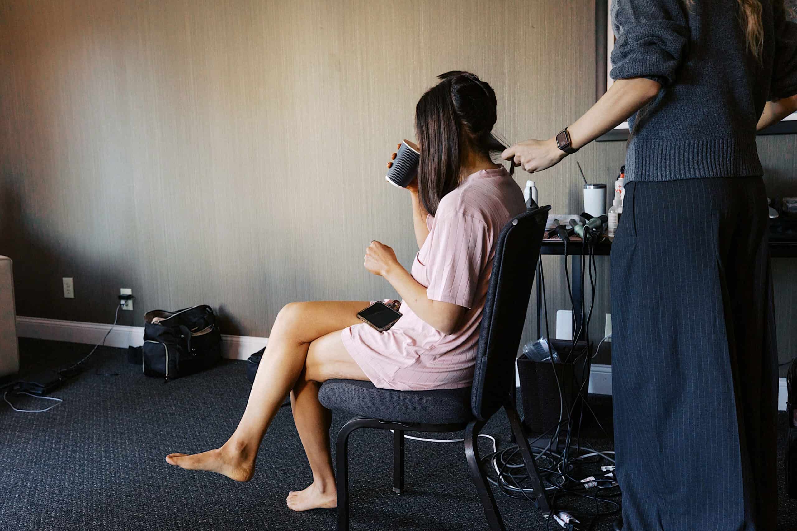 A person in a pink robe sits on a chair, sipping from a mug, while another styles their hair with a tool—capturing the behind-the-scenes elegance of a Philadelphia Cescaphe Wedding at Tendenza amid various hair styling equipment.