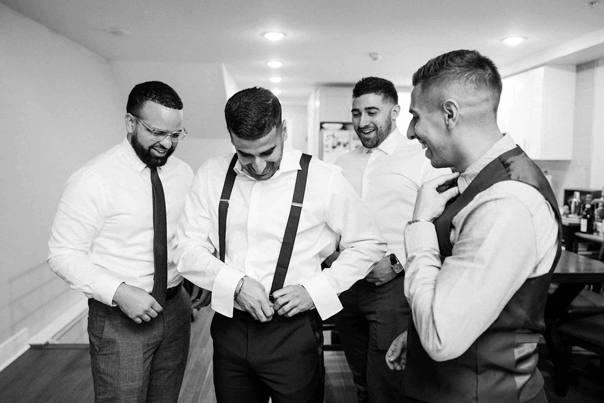 Four men in dress shirts and slacks stand together indoors; one is adjusting his suspenders while the others look on and smile at this elegant Philadelphia Cescaphe Wedding at Tendenza.