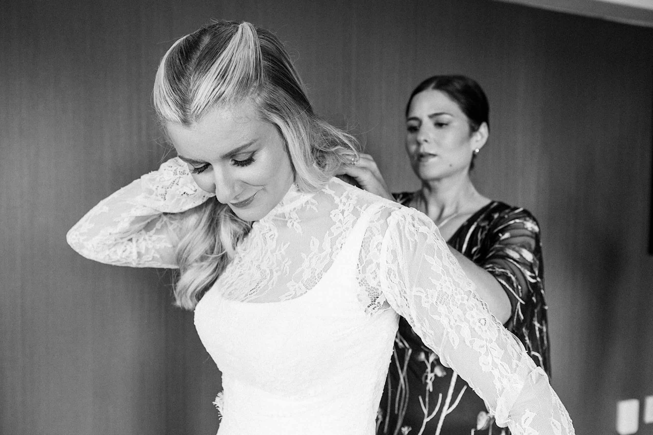 A woman in a lace wedding dress has her gown adjusted by another woman standing behind her, capturing an elegant Philadelphia Cescaphe Wedding at Tendenza.
