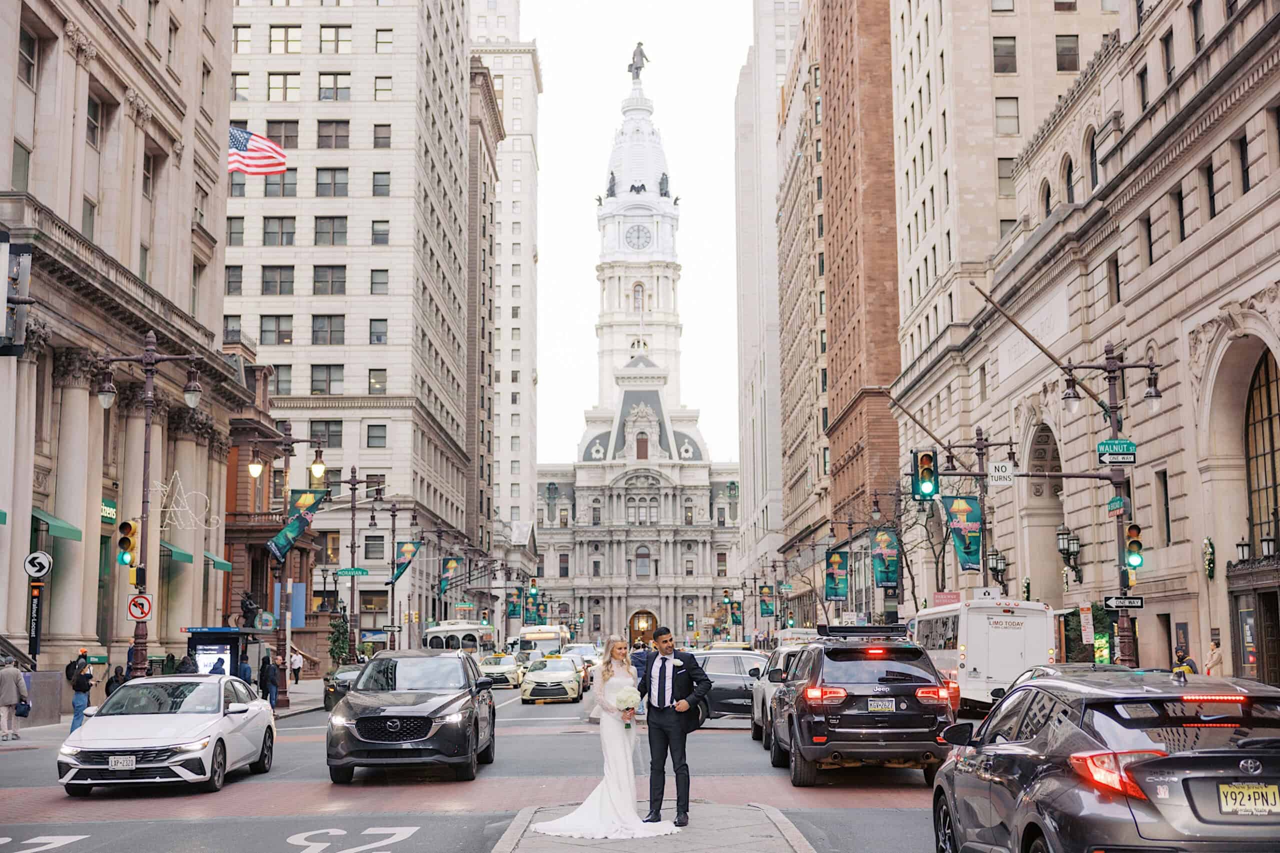 A bride and groom stand in the middle of a busy city street with Philadelphia’s City Hall in the background, capturing an elegant Philadelphia Cescaphe Wedding at Tendenza amid tall buildings and bustling traffic.