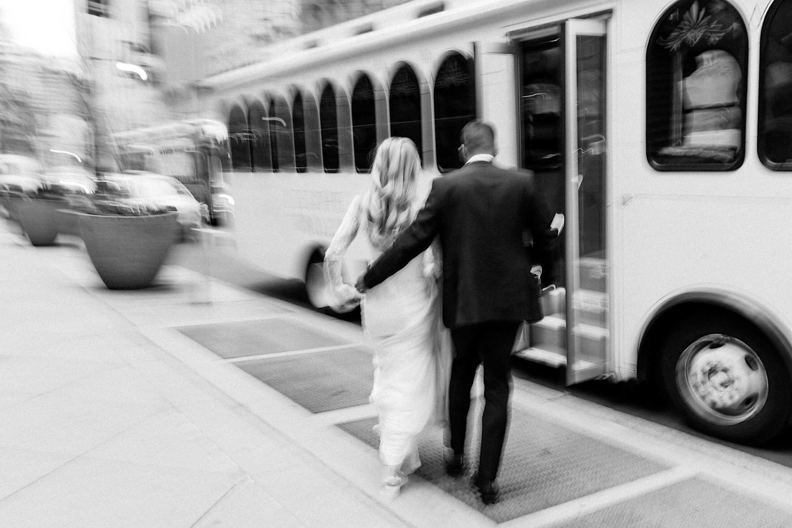 A blurred black and white photo of a couple in formal attire walking towards an open-door trolley bus on a city sidewalk, capturing the elegant charm of a Philadelphia Cescaphe Wedding at Tendenza.