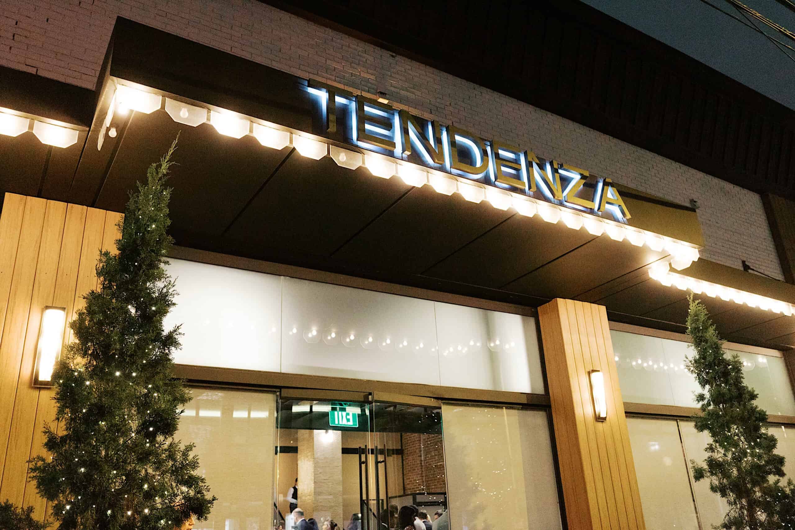 The entrance of Tendenza sets the stage for an elegant Philadelphia Cescaphe Wedding, featuring illuminated signage, glass doors, and two small trees adorned with lights on either side.