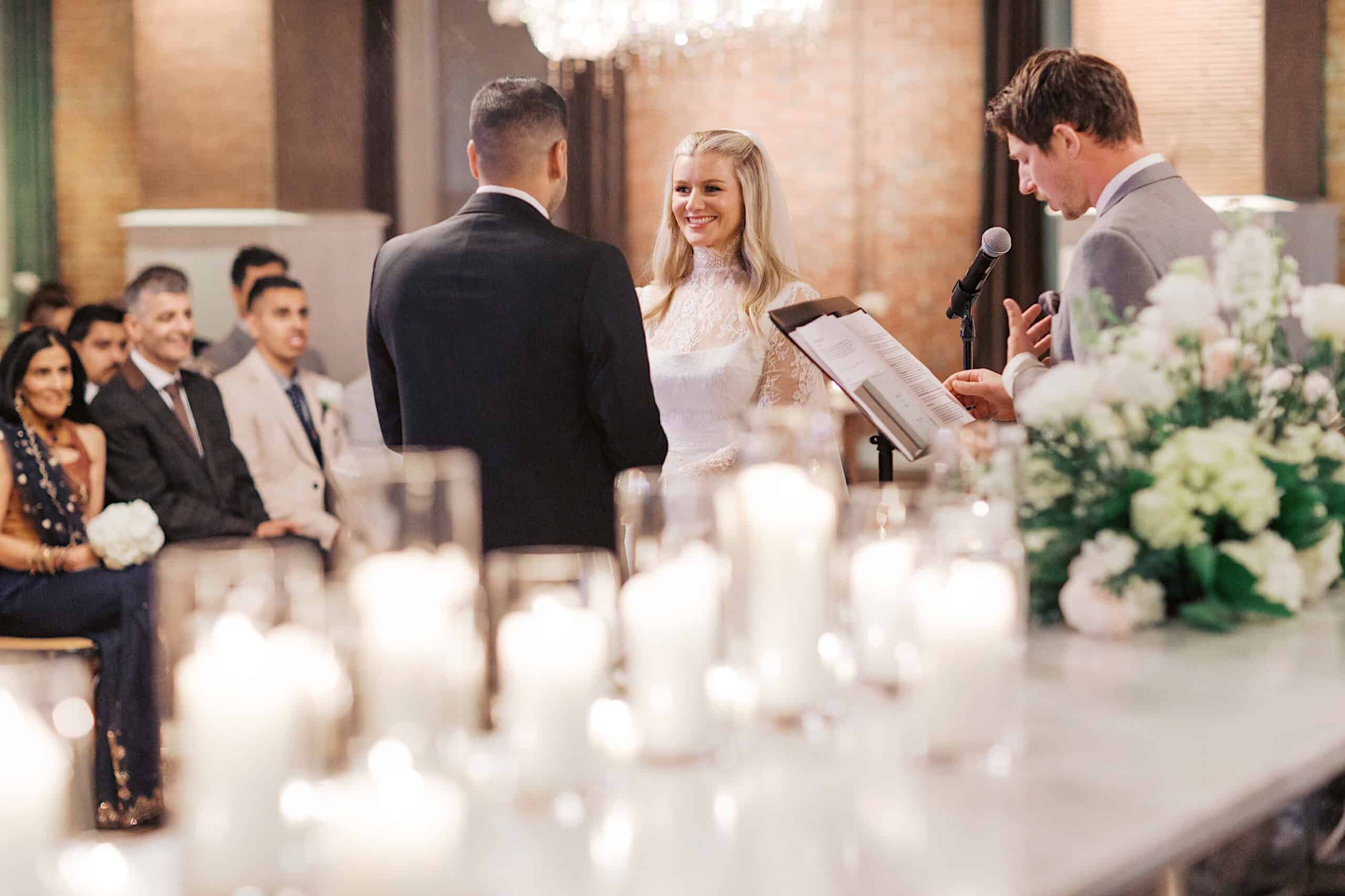 A bride and groom stand together at the altar during their elegant Philadelphia Cescaphe Wedding at Tendenza, as a man officiates and guests look on, with candles softly illuminating the scene.