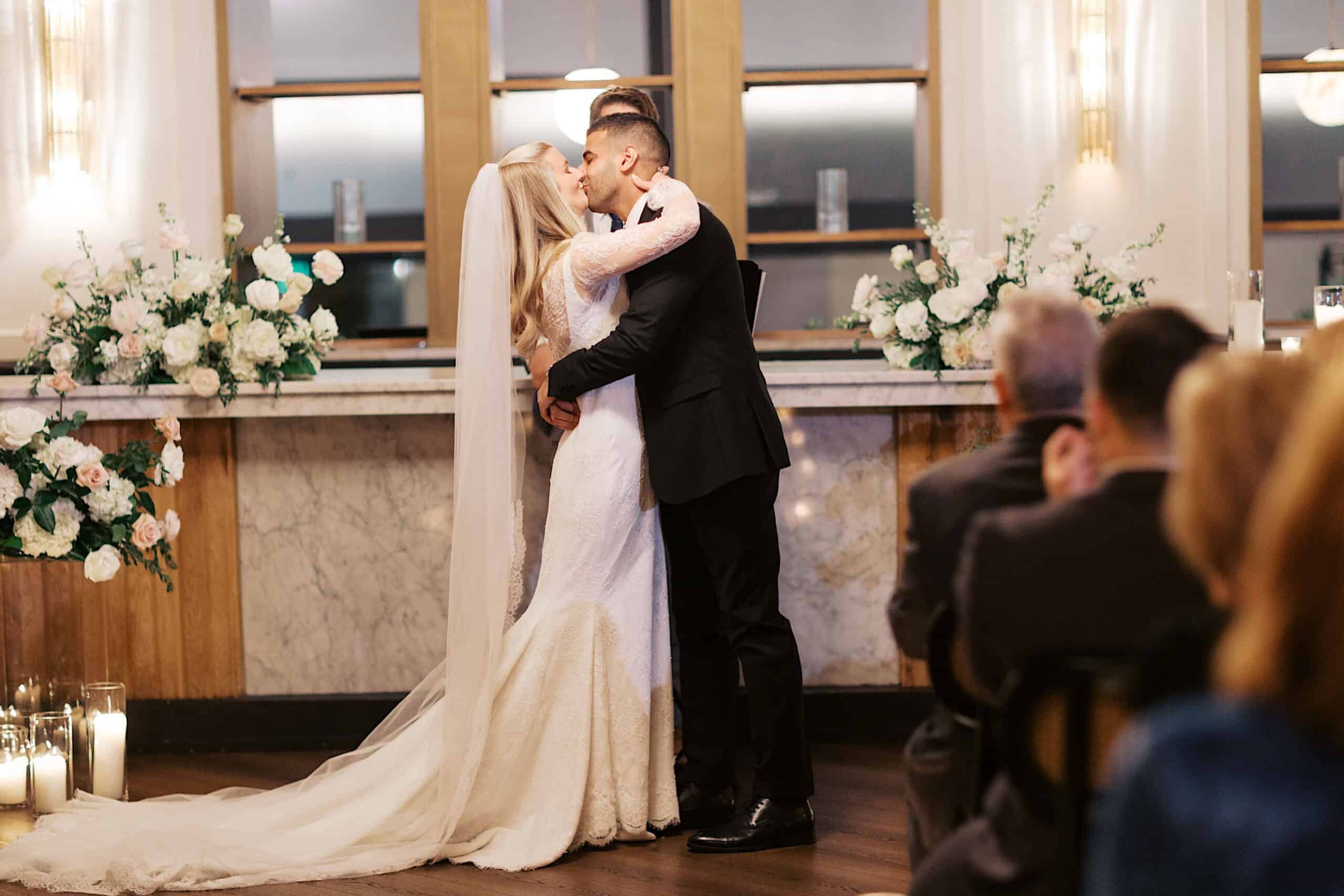 A bride and groom share their first kiss at an elegant Philadelphia Cescaphe Wedding at Tendenza, surrounded by floral arrangements and candles, with guests seated in the foreground.