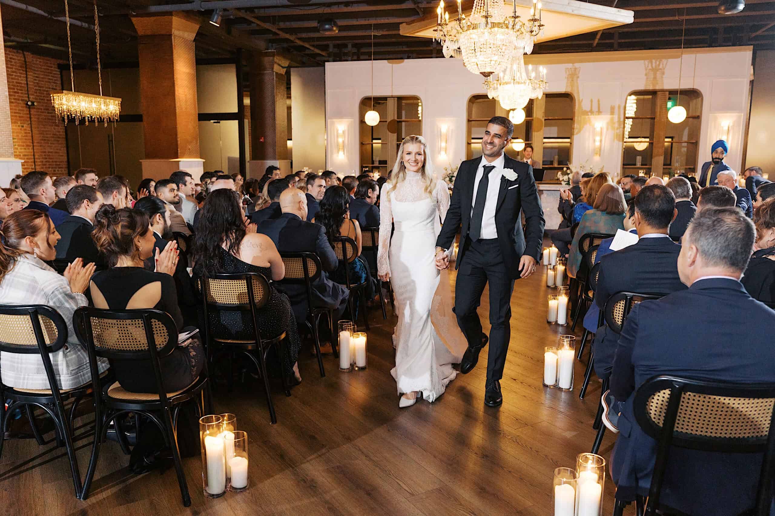 A bride and groom walk down the aisle together in a well-lit indoor venue at Tendenza, surrounded by seated guests and candles lining the floor—an elegant Philadelphia Cescaphe Wedding to remember.