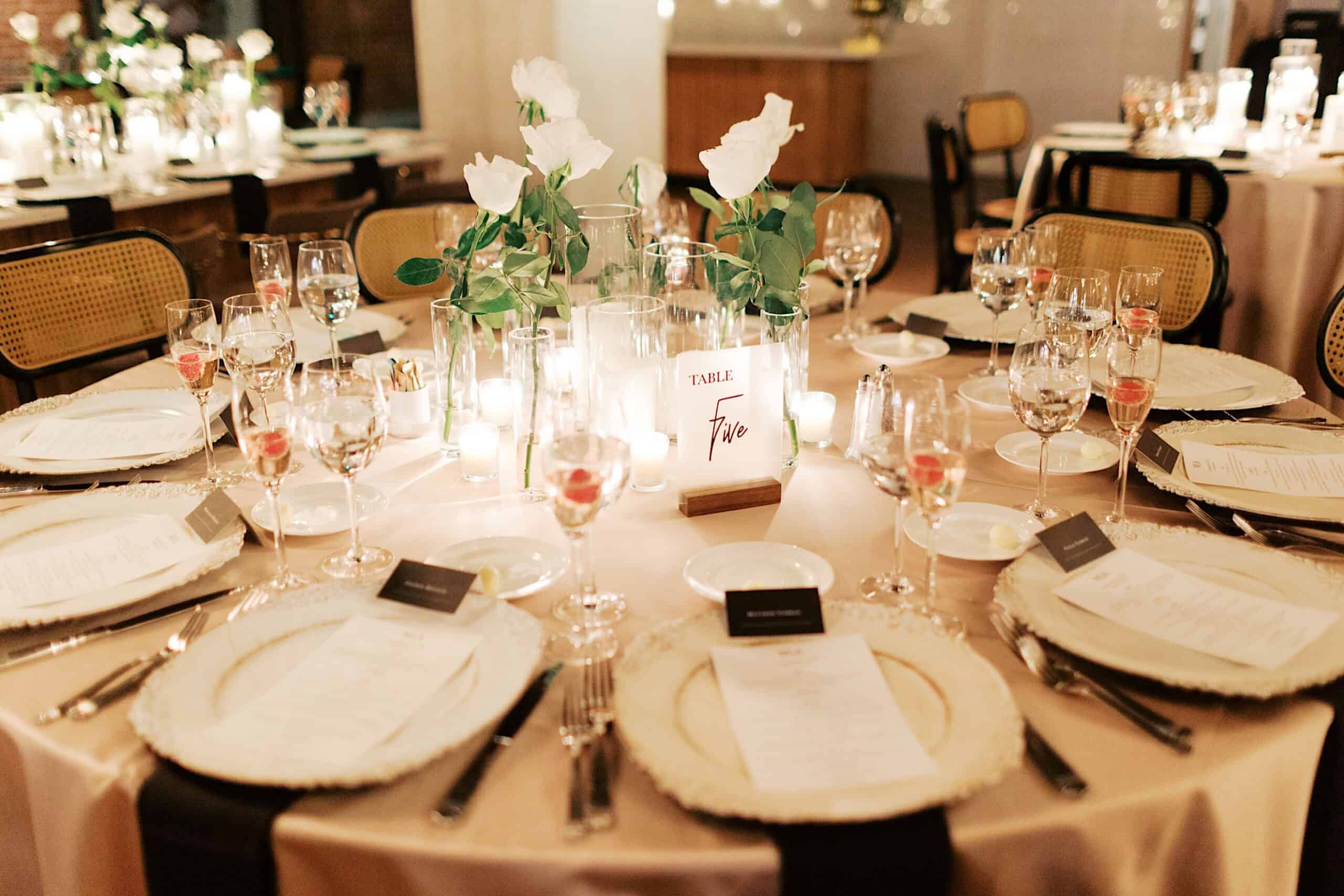 Elegant round dining table set for an event with white plates, menus, glassware, candles, white roses in vases, and a "Table Two" sign at the center—perfect for an elegant Philadelphia Cescaphe Wedding at Tendenza.