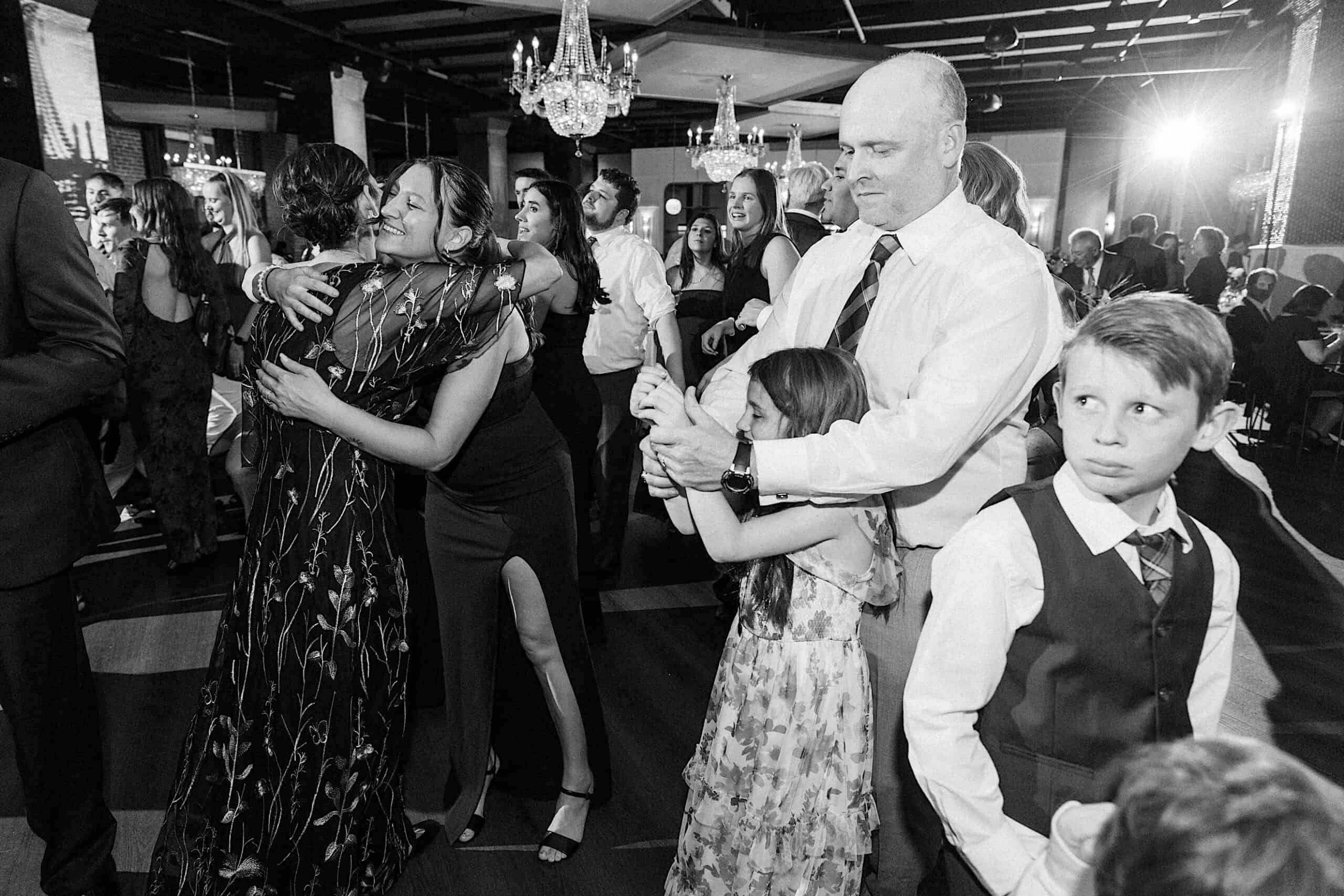 Black-and-white photo of people at an elegant Philadelphia Cescaphe Wedding at Tendenza; some are hugging, others dancing, while children stand nearby among a crowd of adults.