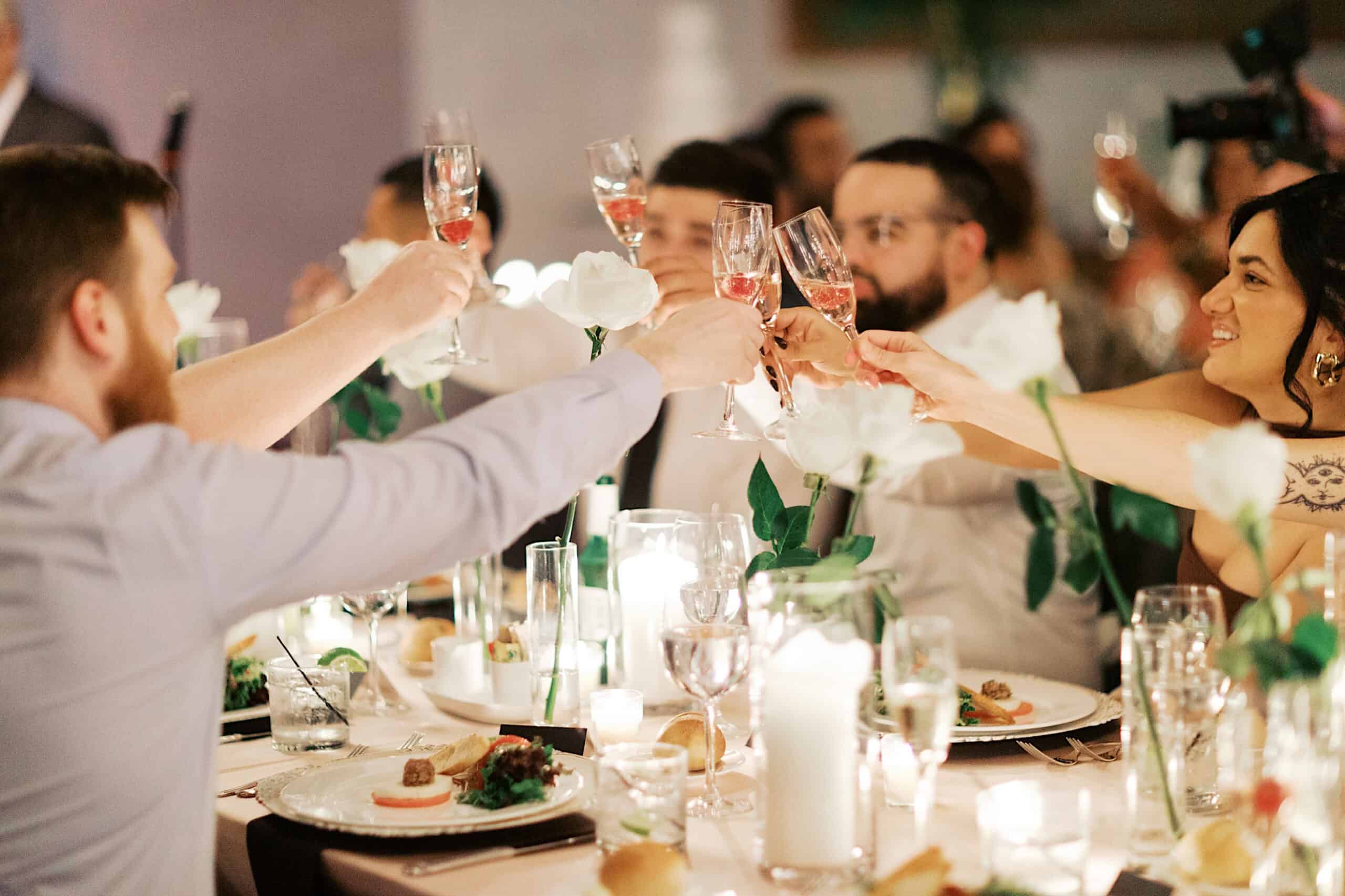 A group of people seated at a decorated table raise glasses in a toast during an elegant Philadelphia Cescaphe Wedding at Tendenza, surrounded by white flowers and candles.