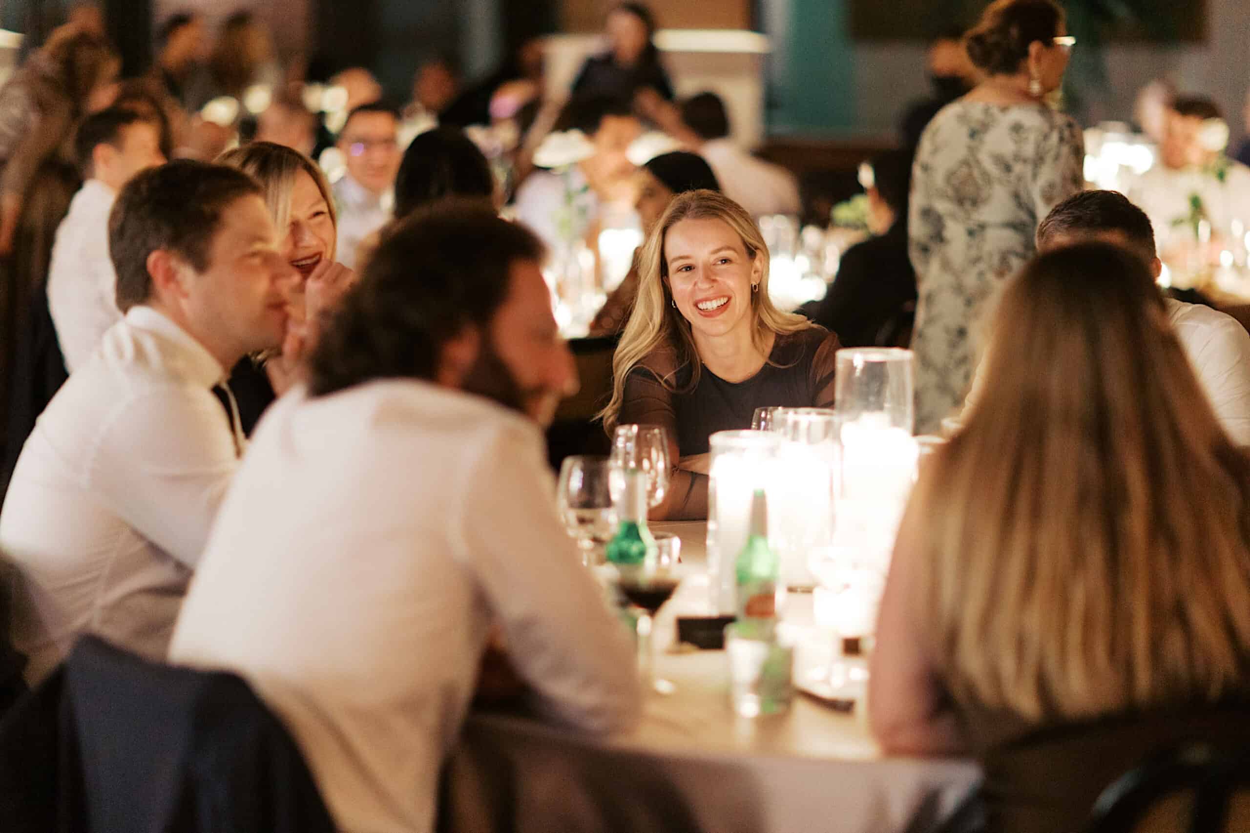 A group of people sit around a candlelit table, talking and smiling in a lively, crowded room at an elegant Philadelphia Cescaphe Wedding at Tendenza.