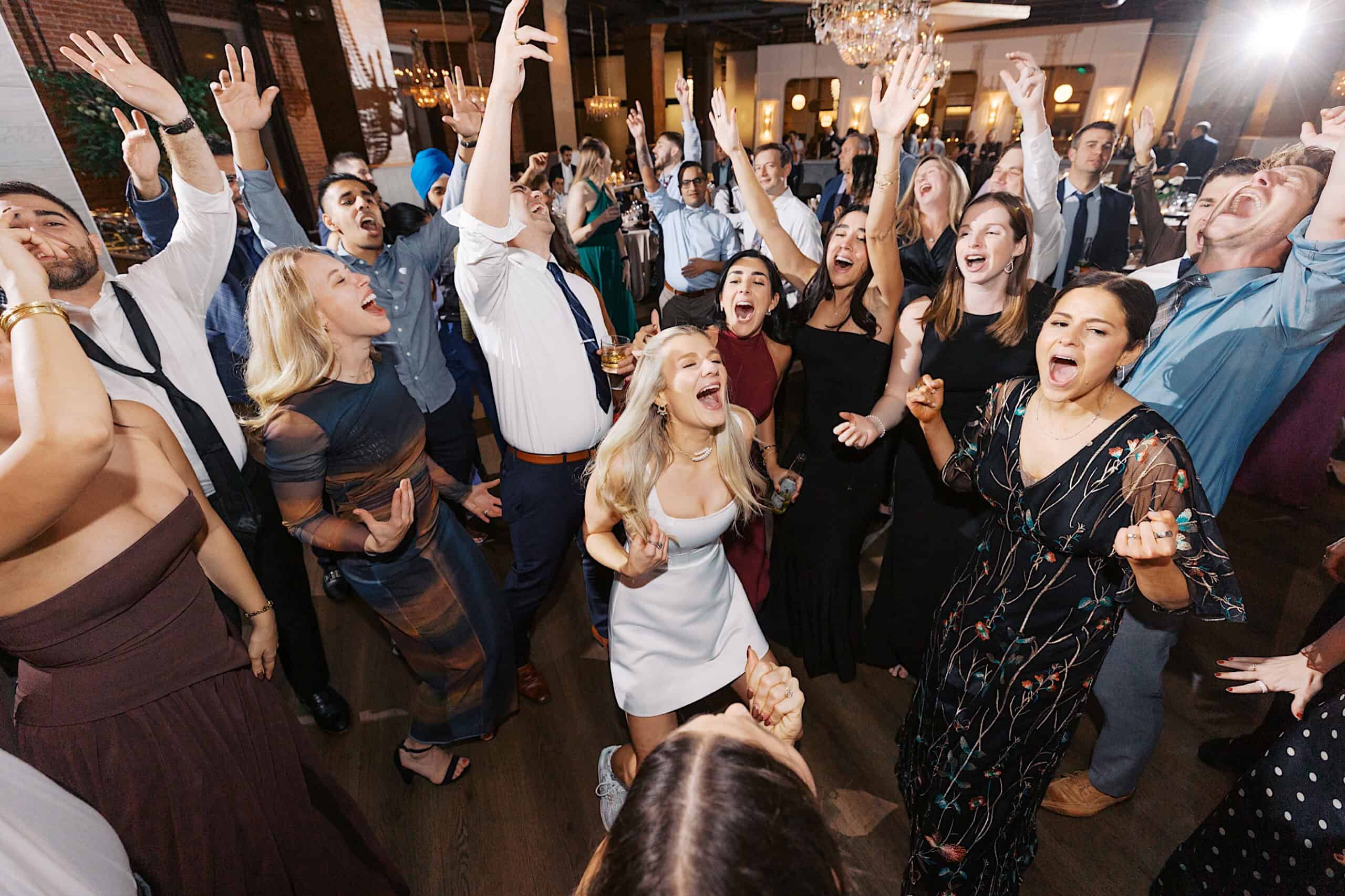 A group of people dressed in formal attire enthusiastically sing and raise their arms on the dance floor during an elegant Philadelphia Cescaphe Wedding at Tendenza.
