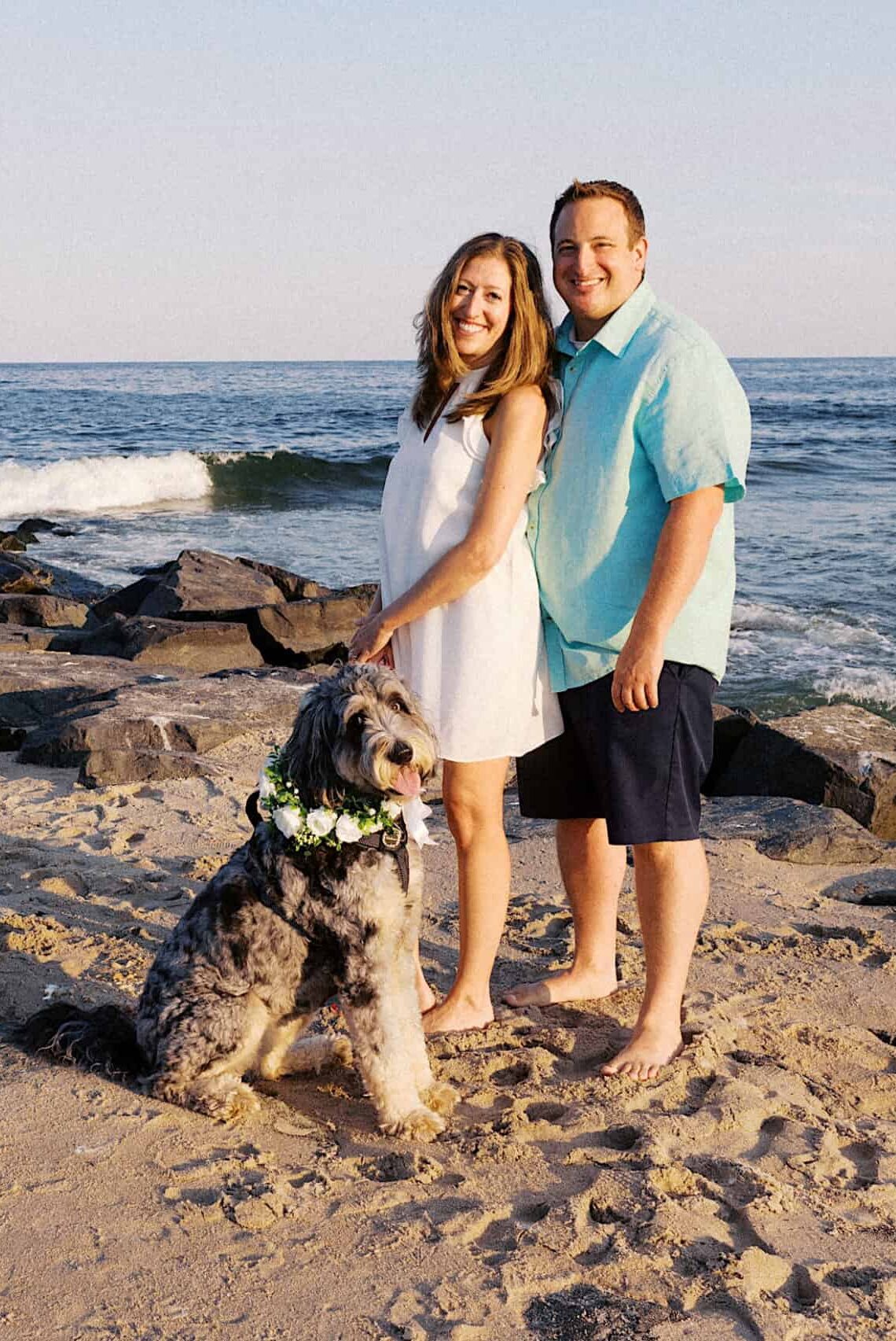 A woman and man stand barefoot on a sandy beach with their dog wearing a flower collar, rocky jetty and ocean waves in the background—capturing the joy of Asbury Park Engagement Photos.