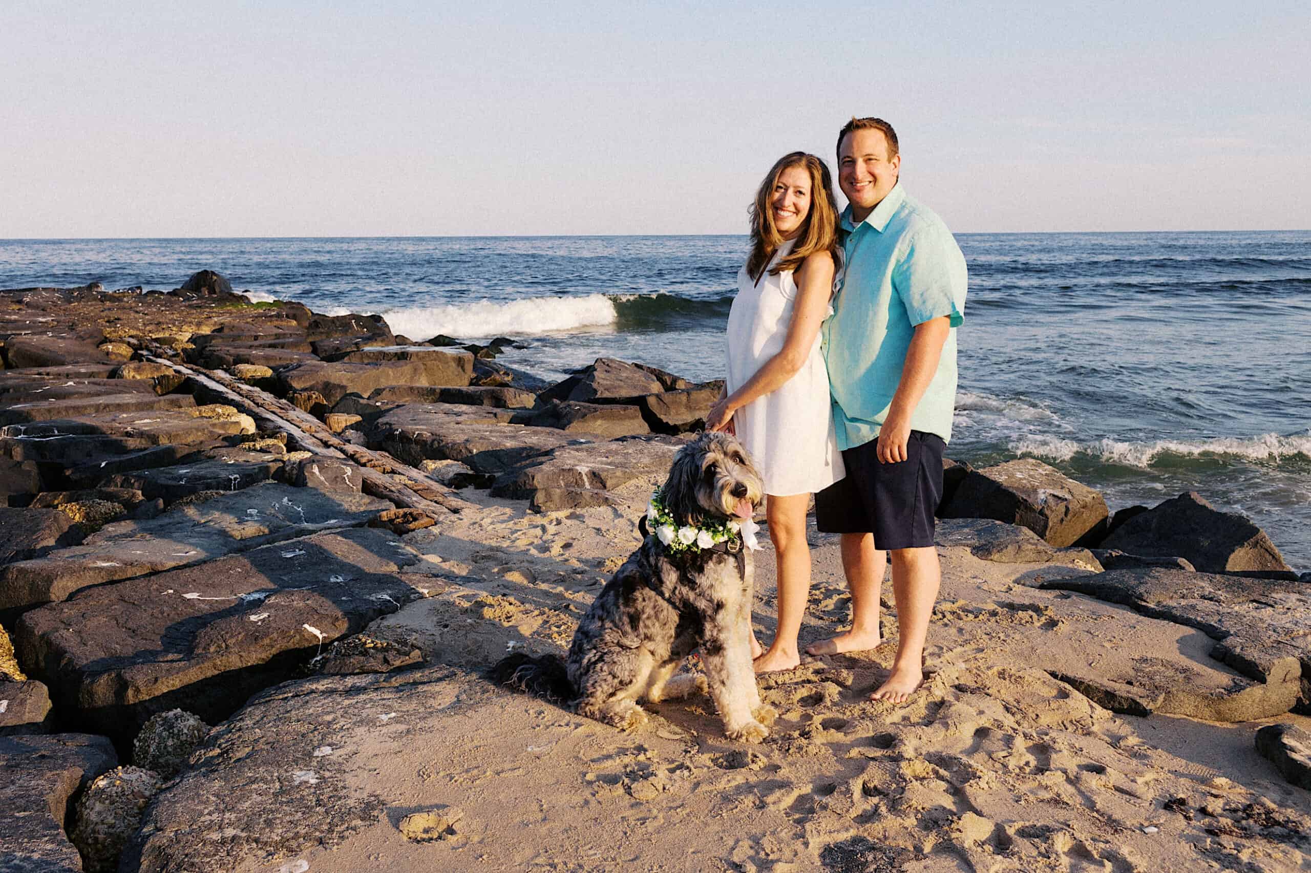 A woman and man stand barefoot on a sandy beach with their dog wearing a flower collar, rocky jetty and ocean waves in the background—capturing the joy of Asbury Park Engagement Photos.