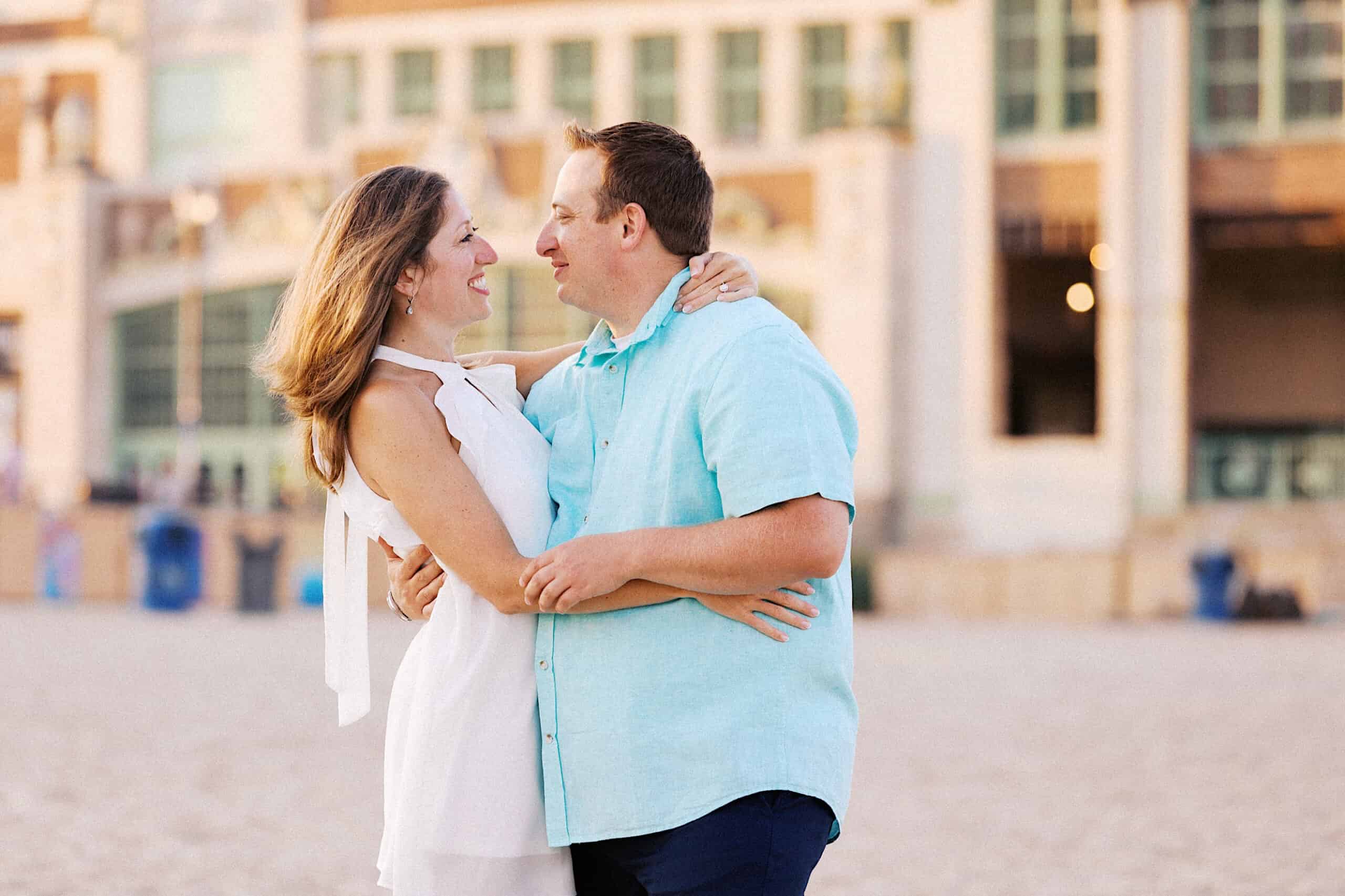 A man and woman stand on a sandy beach embracing and smiling at each other, with a large building in the blurred background—capturing the joy of Asbury Park engagement photos.
