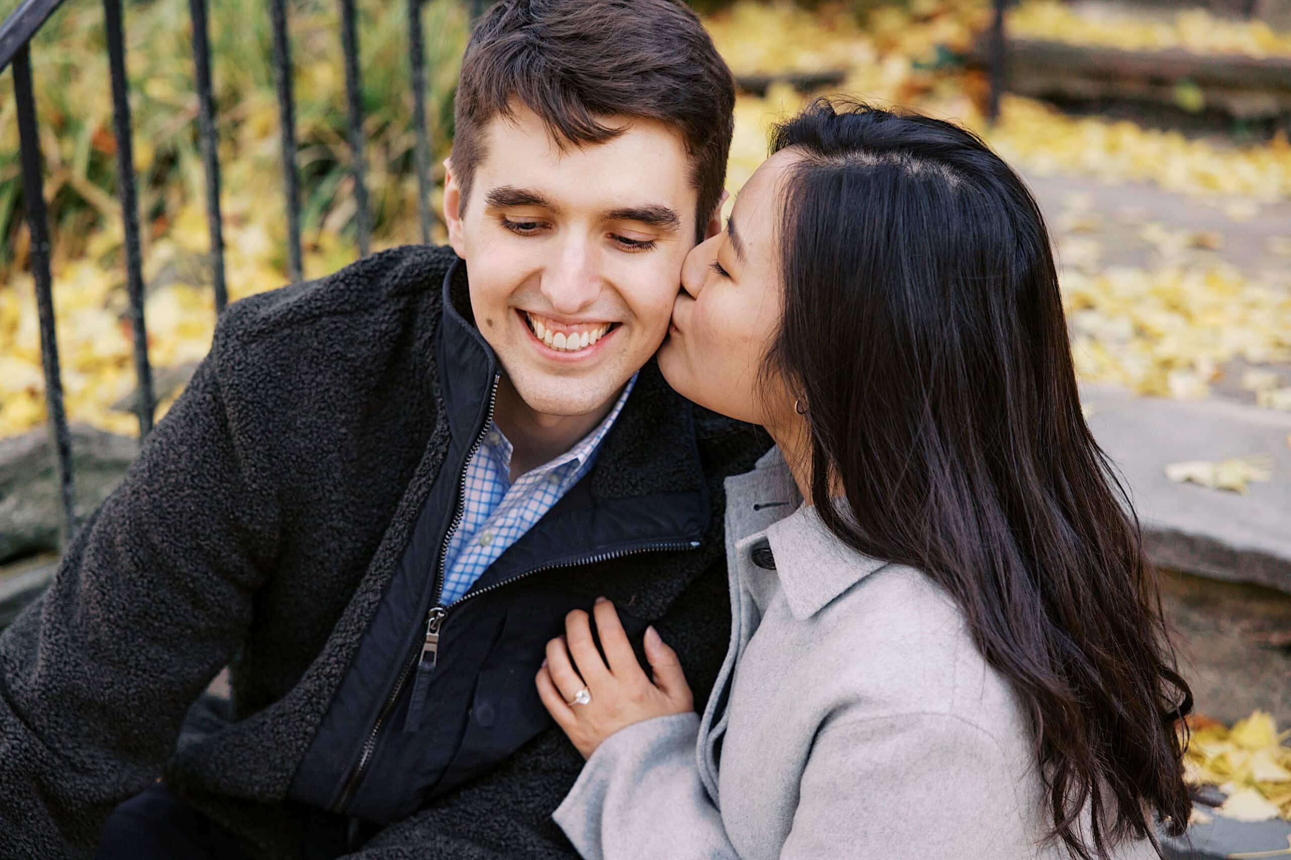 A woman kisses a smiling man on the cheek while sitting on outdoor steps surrounded by autumn leaves, capturing beautiful fall engagement photos in Chestnut Hill.