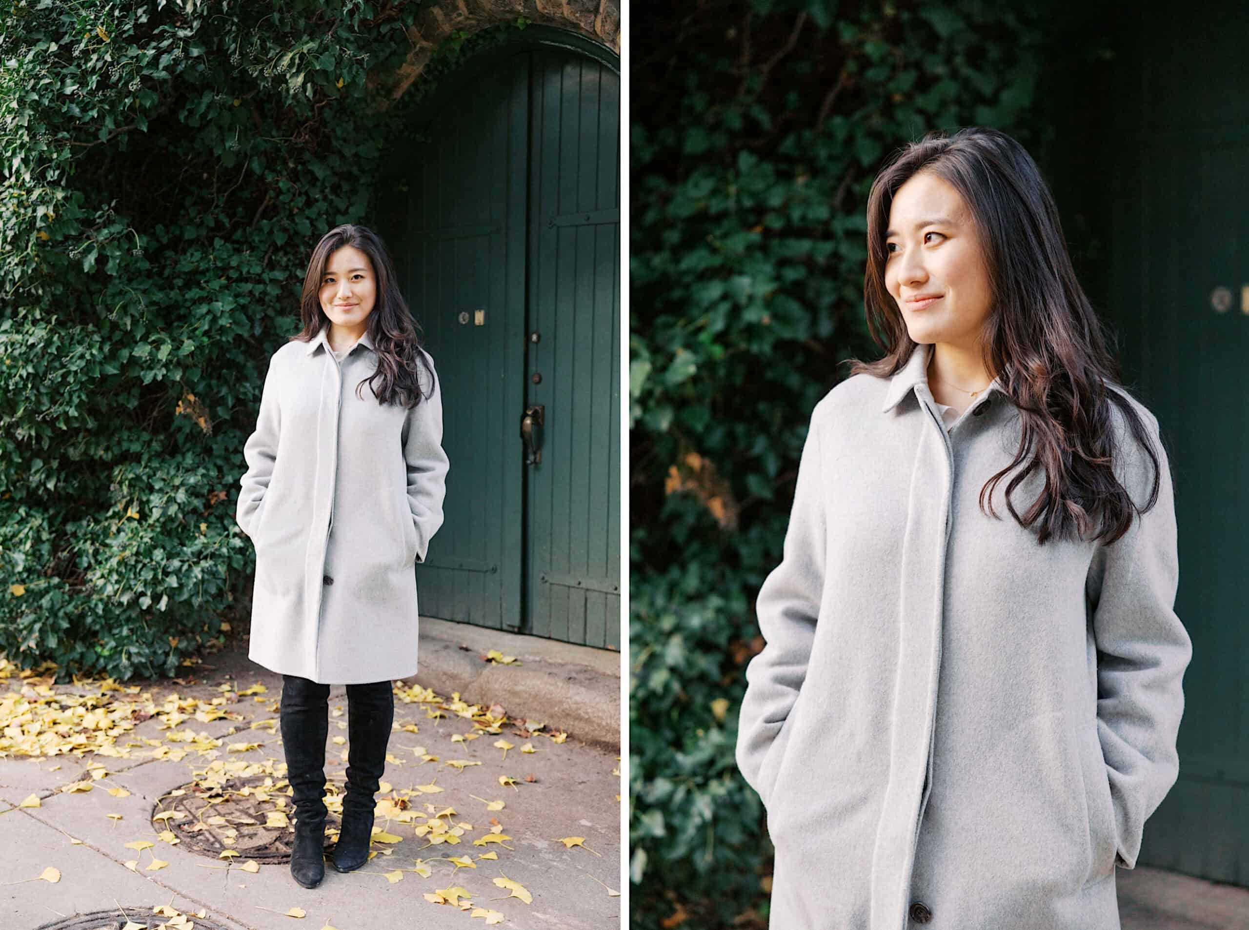 A woman wearing a light gray coat and black boots stands outdoors by a green door and leafy vines, with yellow leaves scattered on the ground—perfect for fall engagement photos in Chestnut Hill.