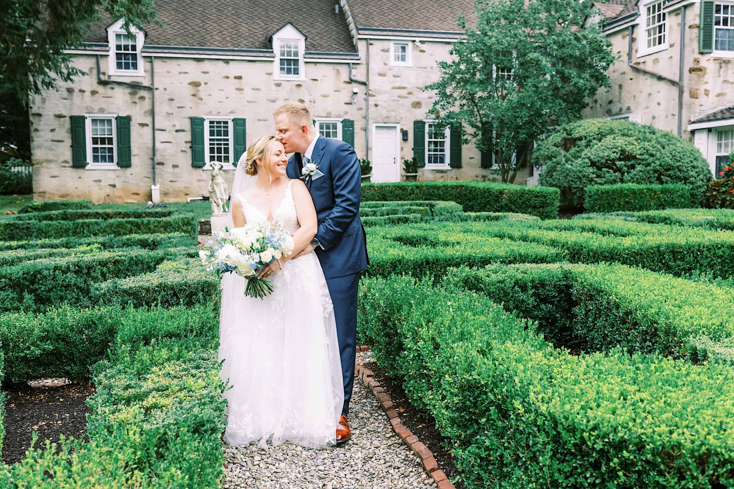 A bride and groom stand closely together in a manicured garden, smiling, during their Summer Wedding at Appleford Estate, with a stone building featuring white-framed windows and green shutters in the background.