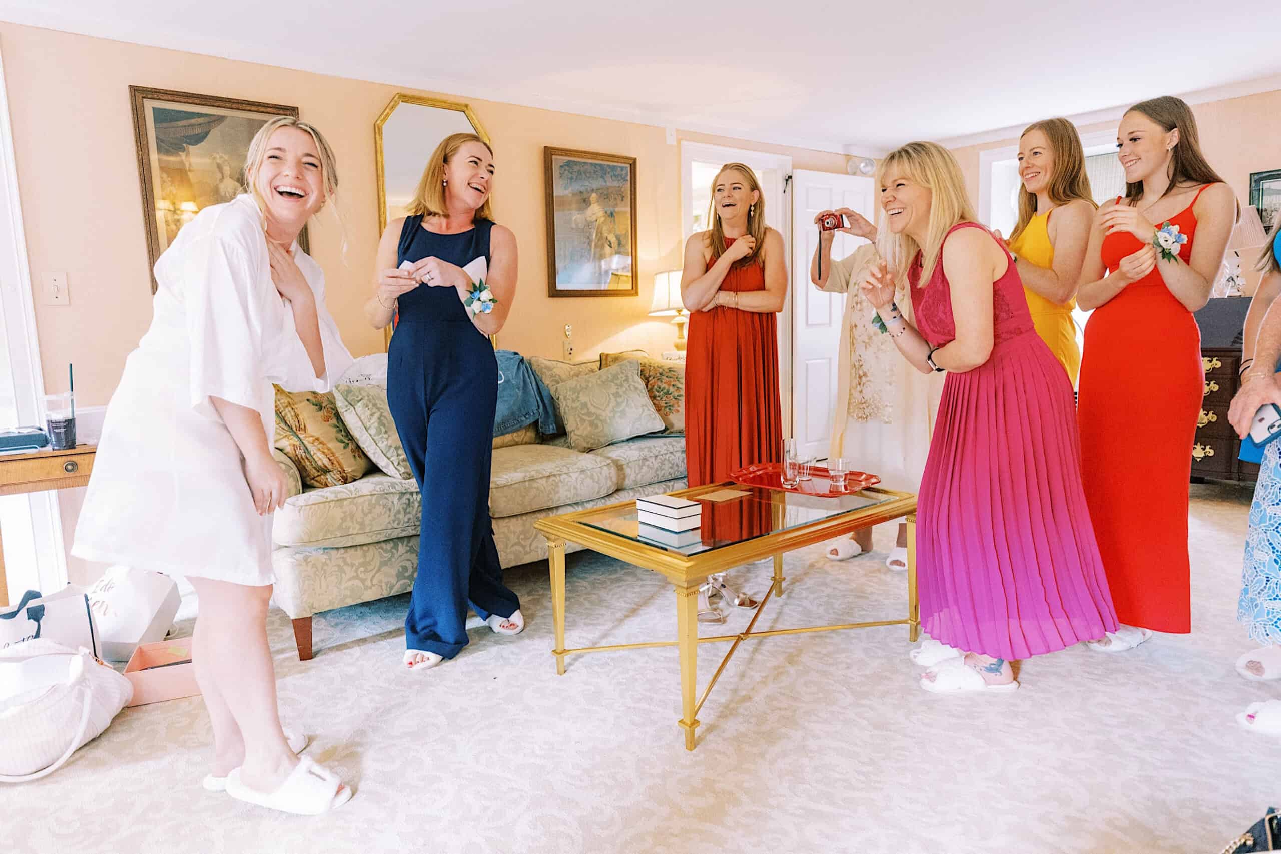 Six women in colorful dresses and slippers stand and laugh together in a brightly lit living room, sharing joyful moments before a Summer Wedding at Appleford Estate. A cozy couch, table, and mirrors complete the charming scene.
