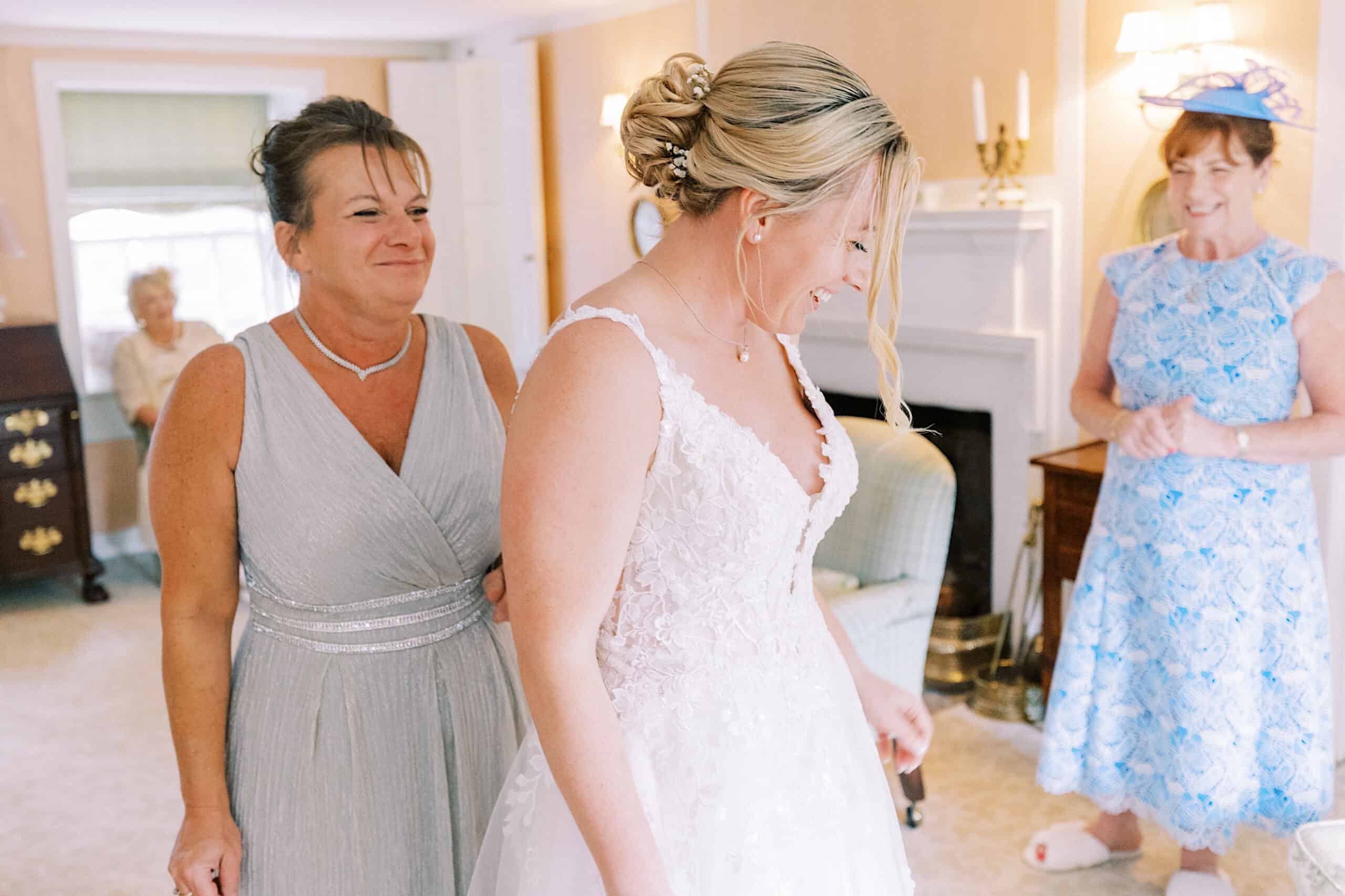 A bride in a white gown smiles while two women in light-colored dresses stand nearby in a softly lit room during a Summer Wedding at Appleford Estate.
