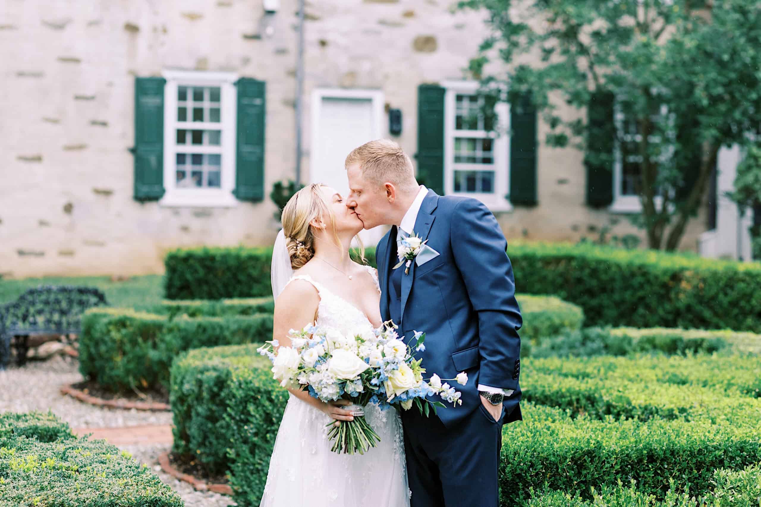 A bride and groom kiss in a garden during their Summer Wedding at Appleford Estate, with the bride holding a bouquet of flowers and a building with green shutters in the background.