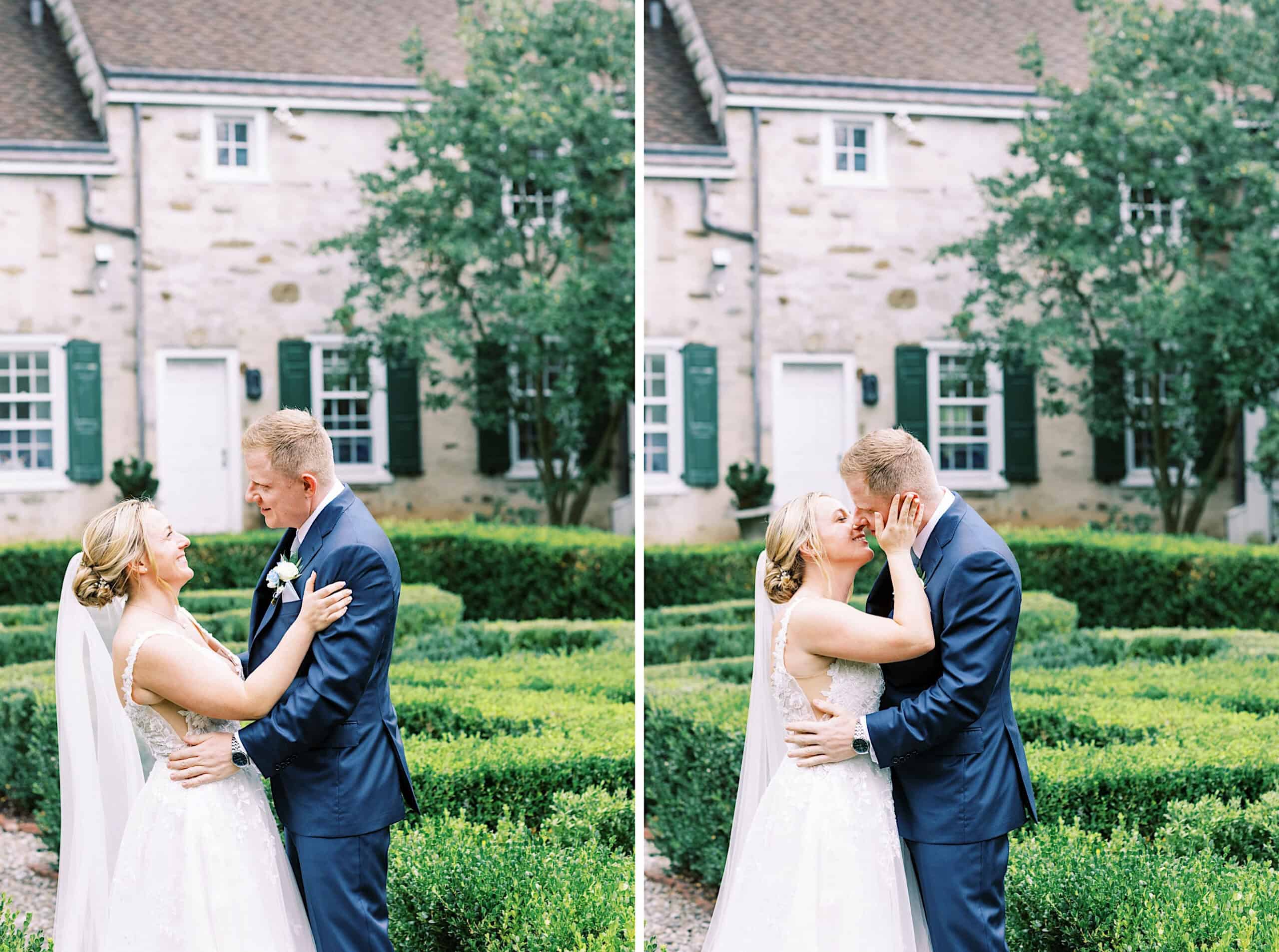 A bride and groom stand together in front of a stone building with trimmed hedges, embracing and sharing a kiss during their summer wedding at Appleford Estate.