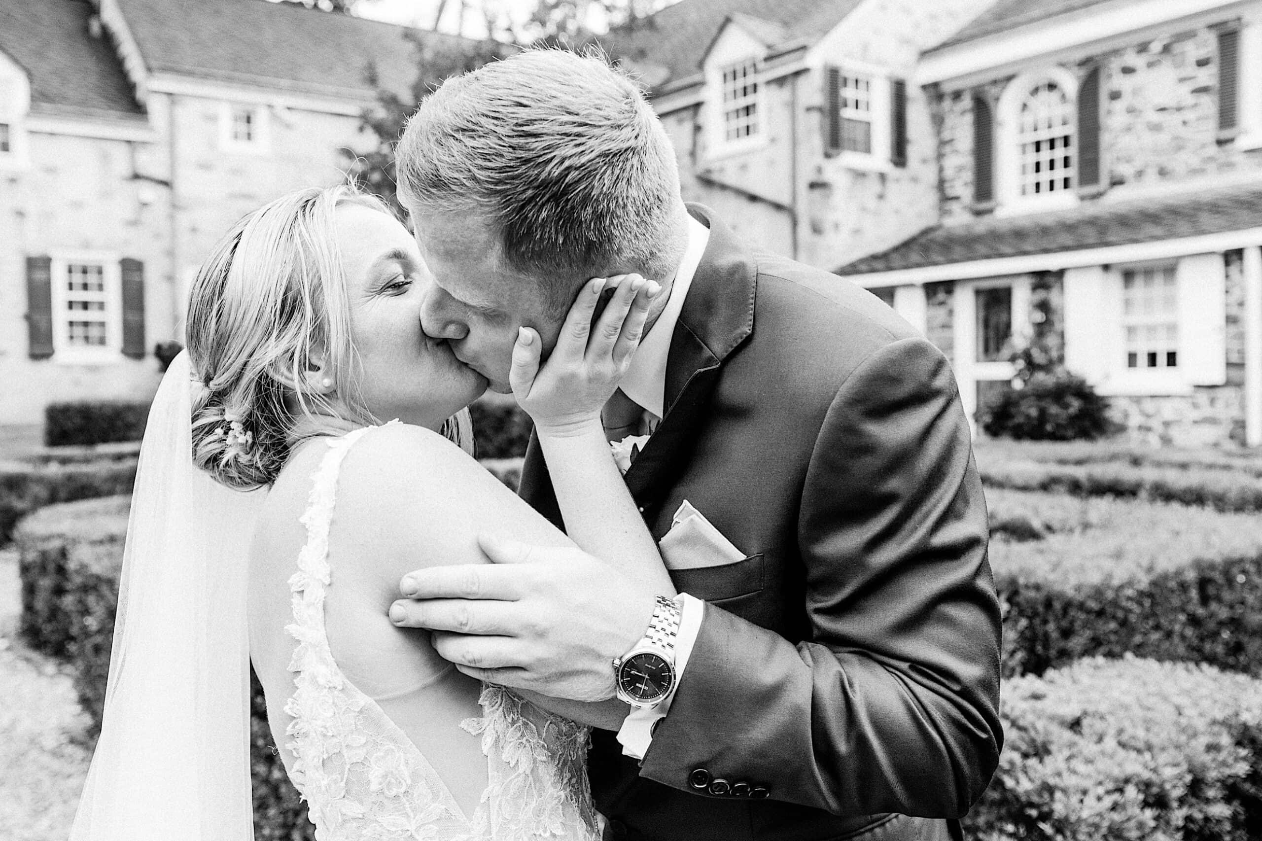 A bride and groom share a kiss outside in wedding attire at their Summer Wedding at Appleford Estate, with a historic stone building and garden in the background.