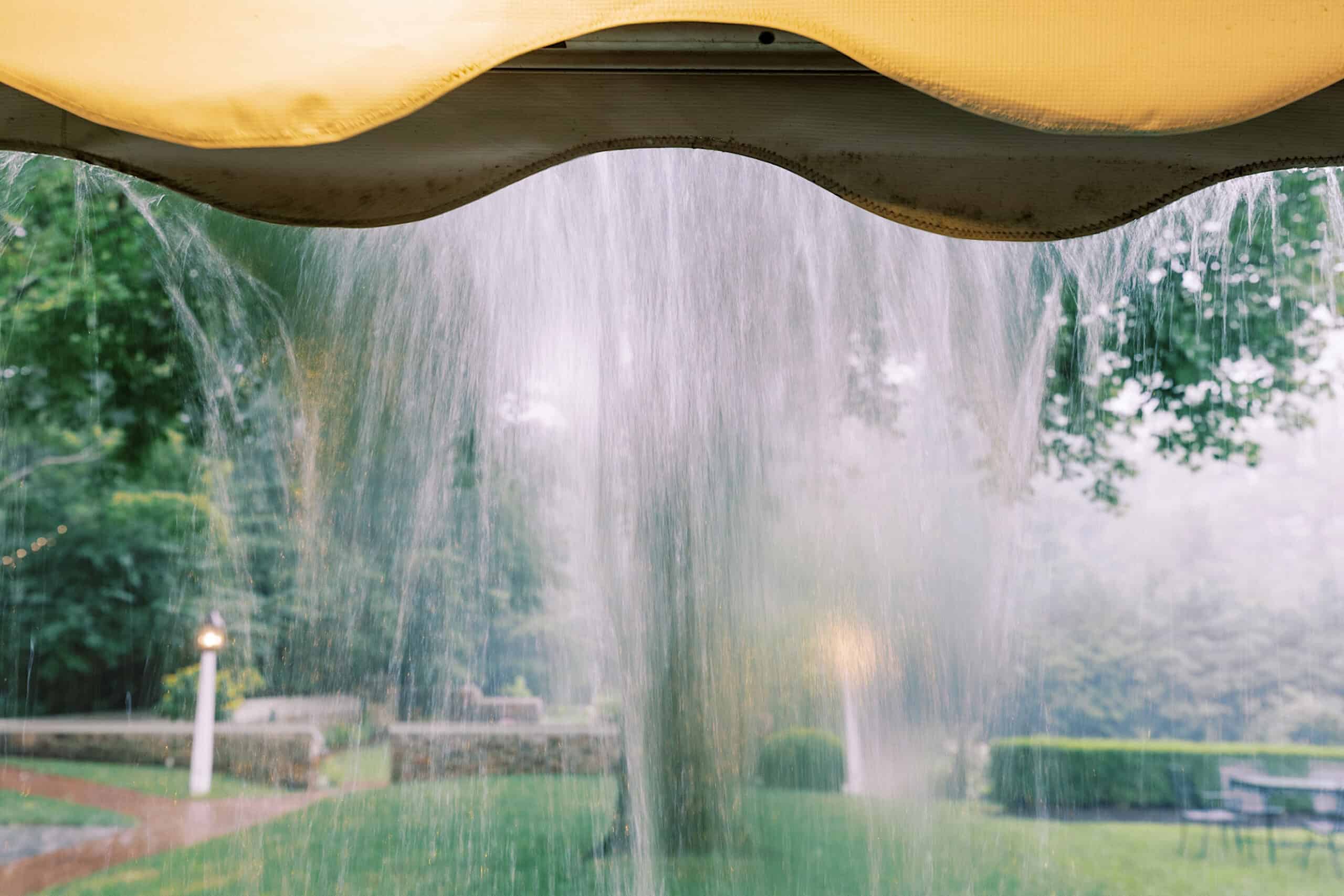 View from under a canopy as rainwater pours off the edge, with a garden and outdoor furniture in the background—an enchanting scene reminiscent of a Summer Wedding at Appleford Estate.
