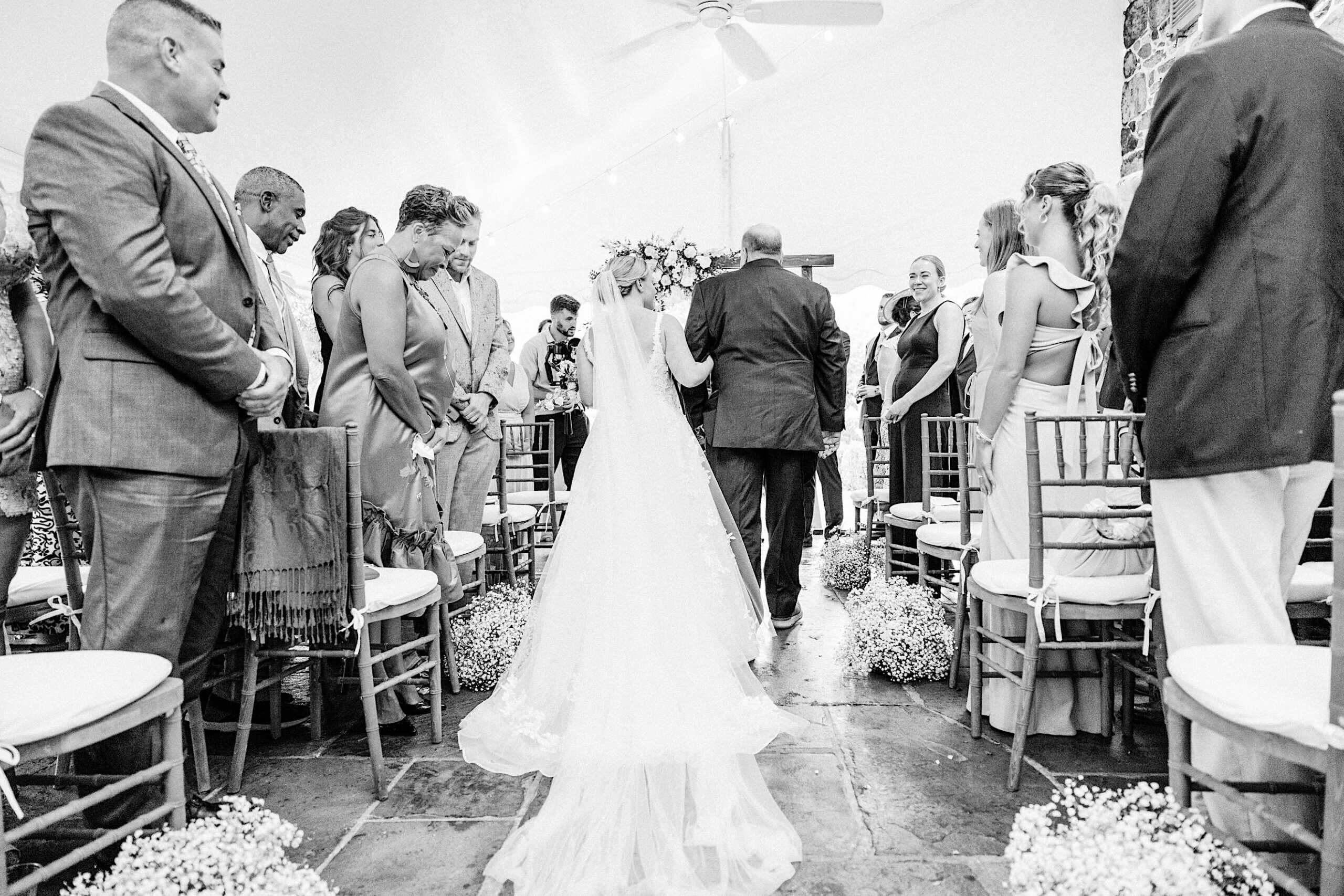 A bride in a wedding dress walks down the aisle with a man, as guests stand on either side, celebrating a Summer Wedding at Appleford Estate indoors under a tent.