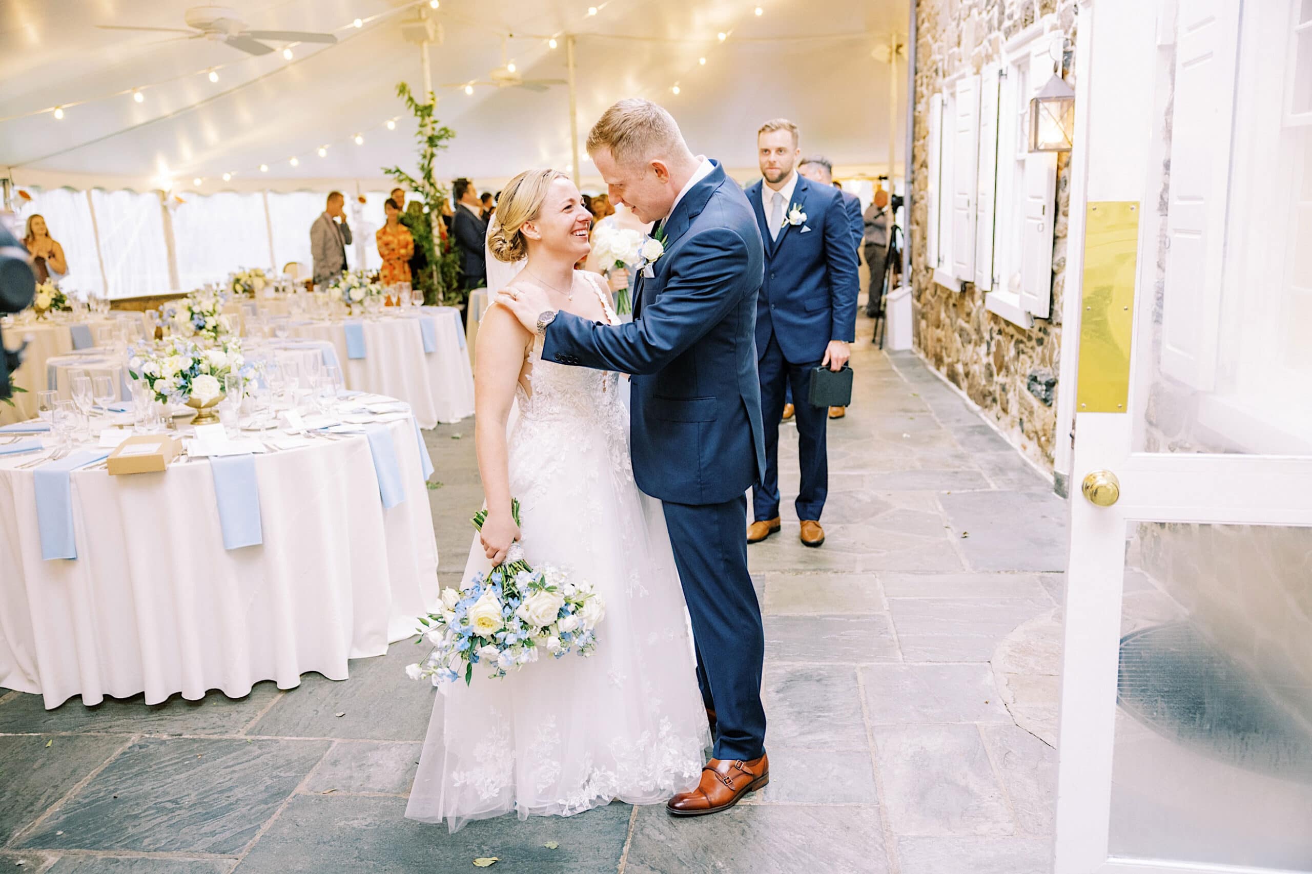 A bride and groom stand close together smiling inside a wedding reception venue with decorated tables and guests in the background, celebrating their Summer Wedding at Appleford Estate.