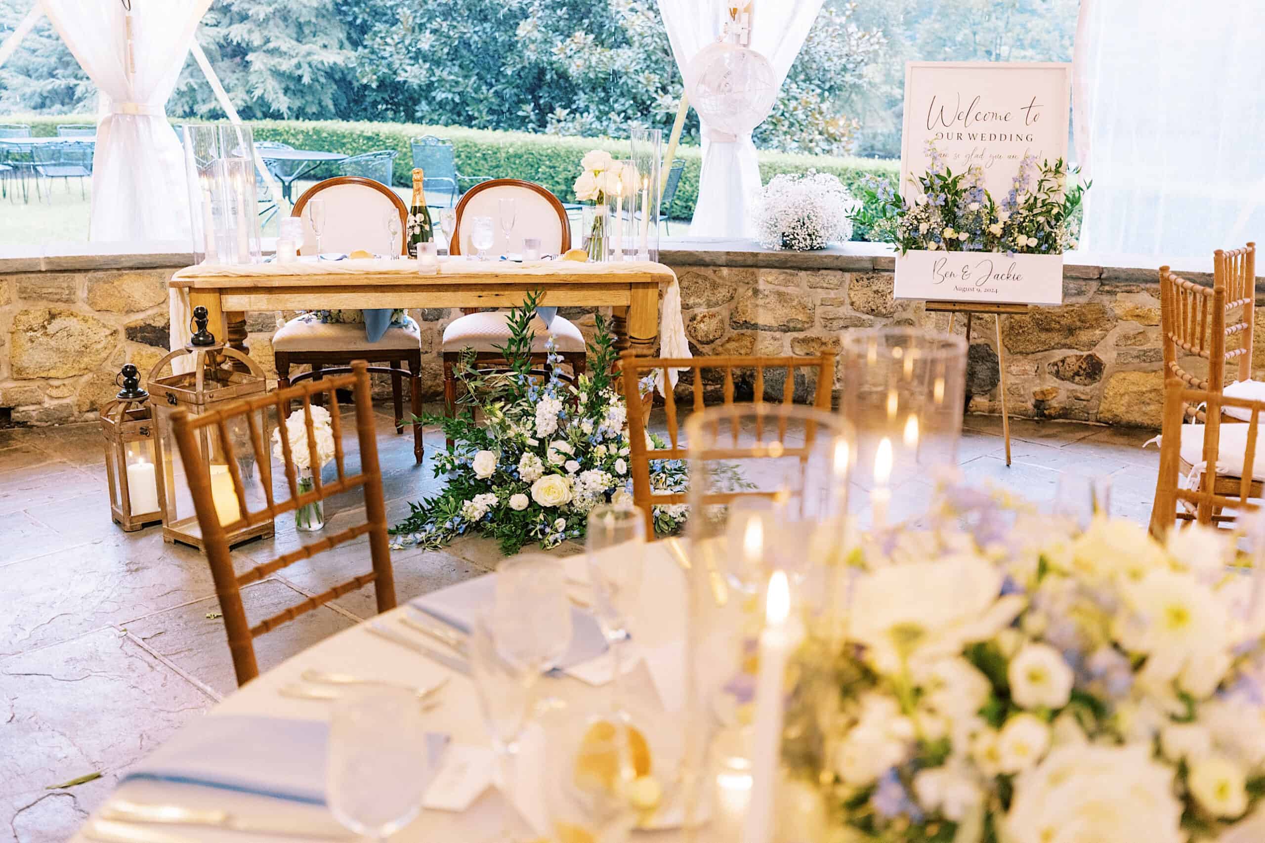 Elegant Summer Wedding at Appleford Estate, featuring decorated tables, lush floral arrangements, wooden chairs, and a charming welcome sign by a stone wall under a graceful tent.