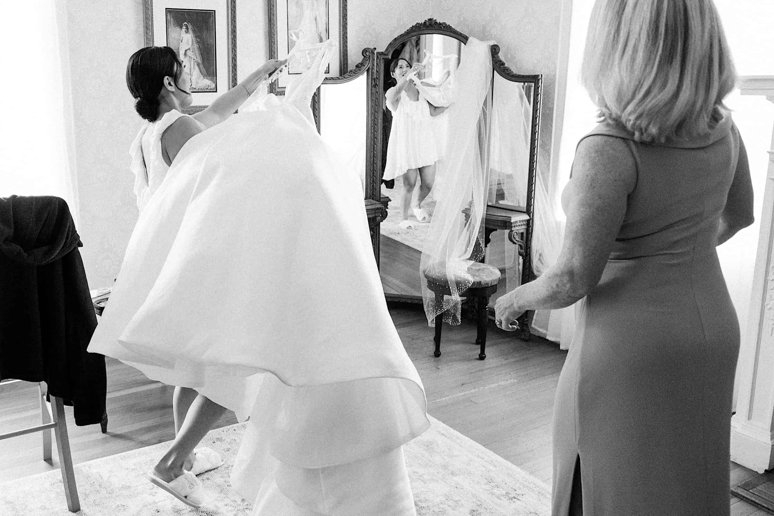 A bride lifts her wedding dress while preparing in front of a mirror, with another woman standing nearby in a bedroom before her elegant Cairnwood Estate Wedding.