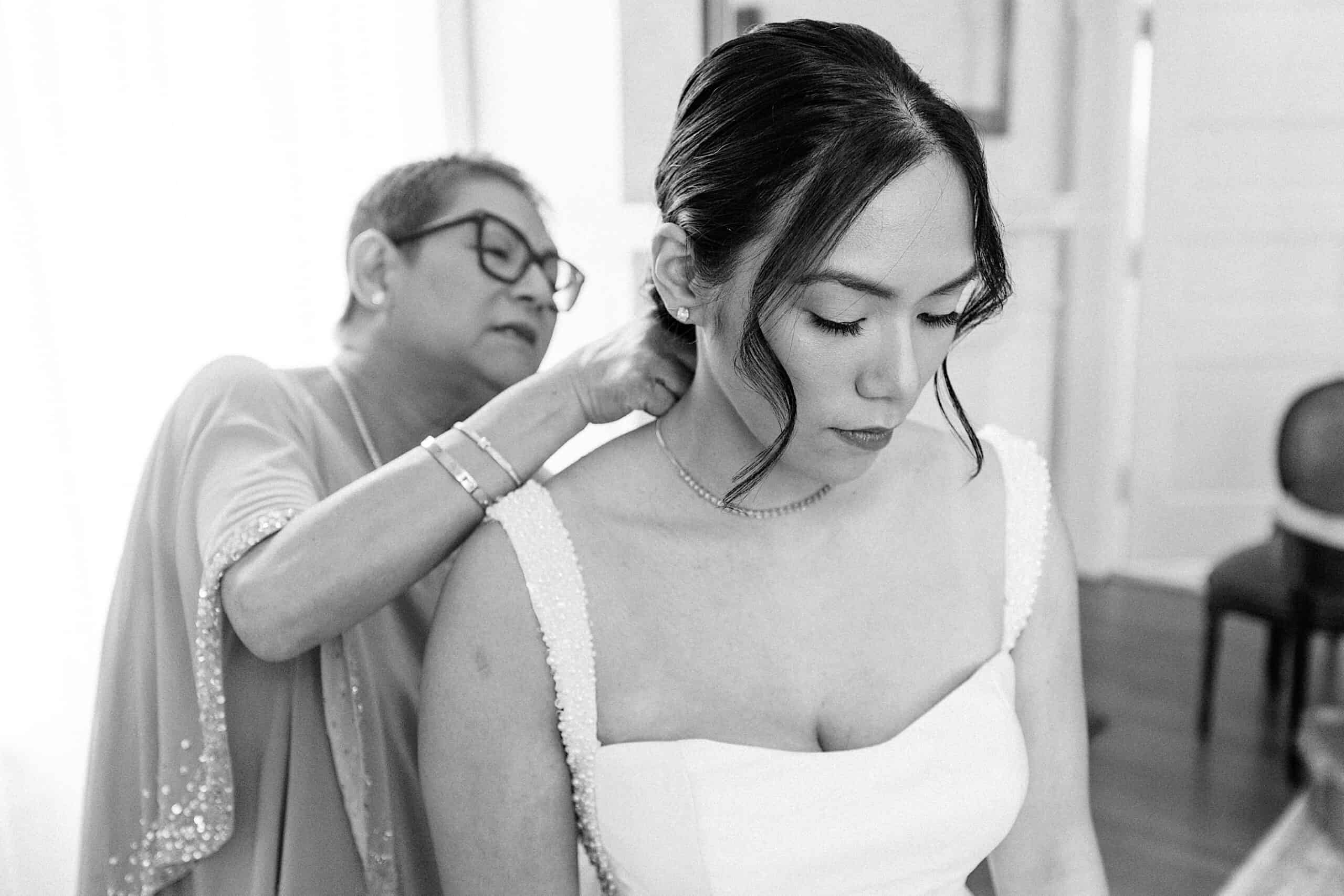 A woman fastens a necklace around the neck of a bride dressed in a white gown, the softly lit room capturing an elegant Cairnwood Estate Wedding moment.