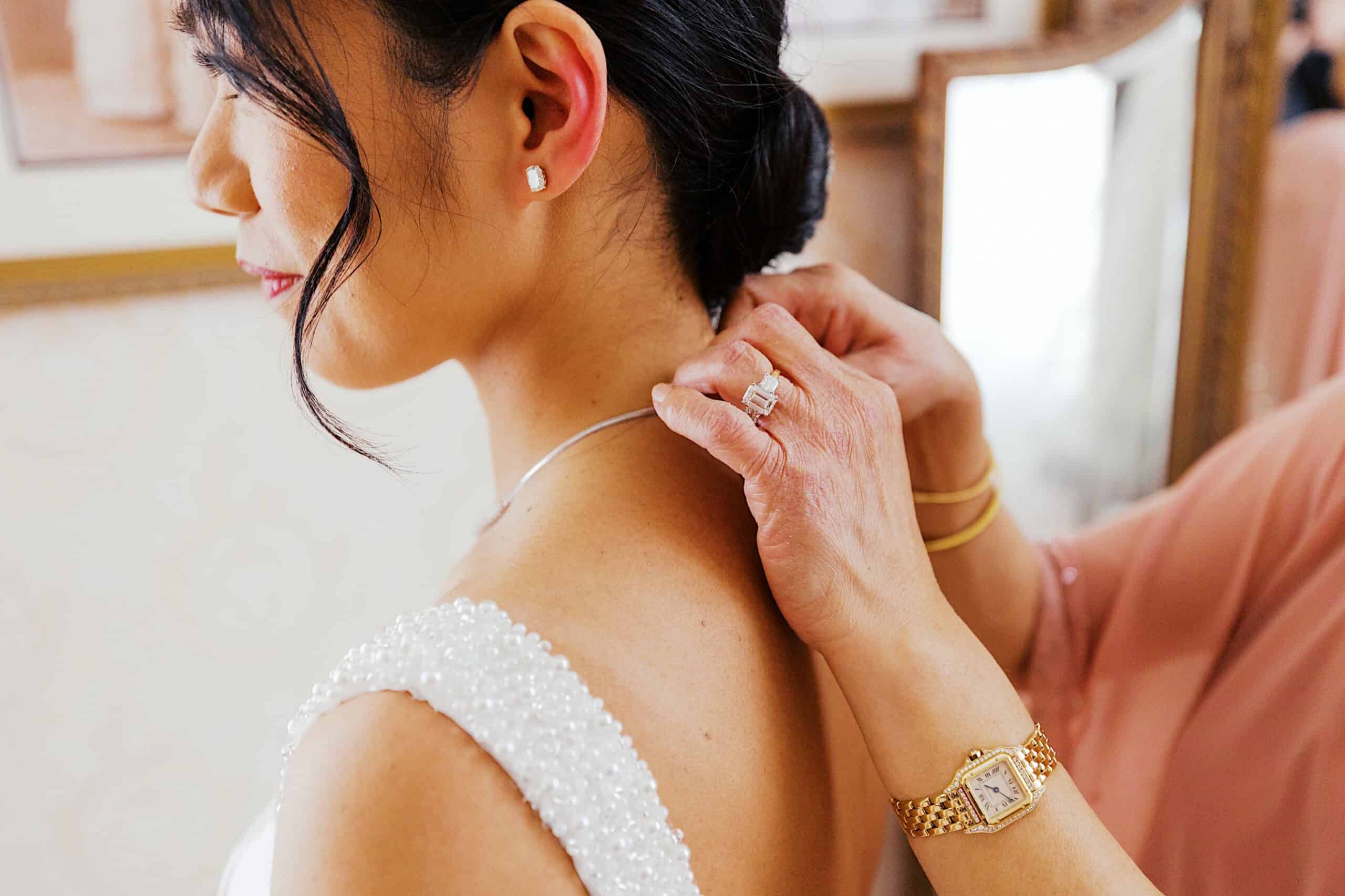A woman fastens a necklace around the neck of a bride in a white beaded dress during an elegant Cairnwood Estate Wedding. The scene is indoors, with framed pictures in the background.