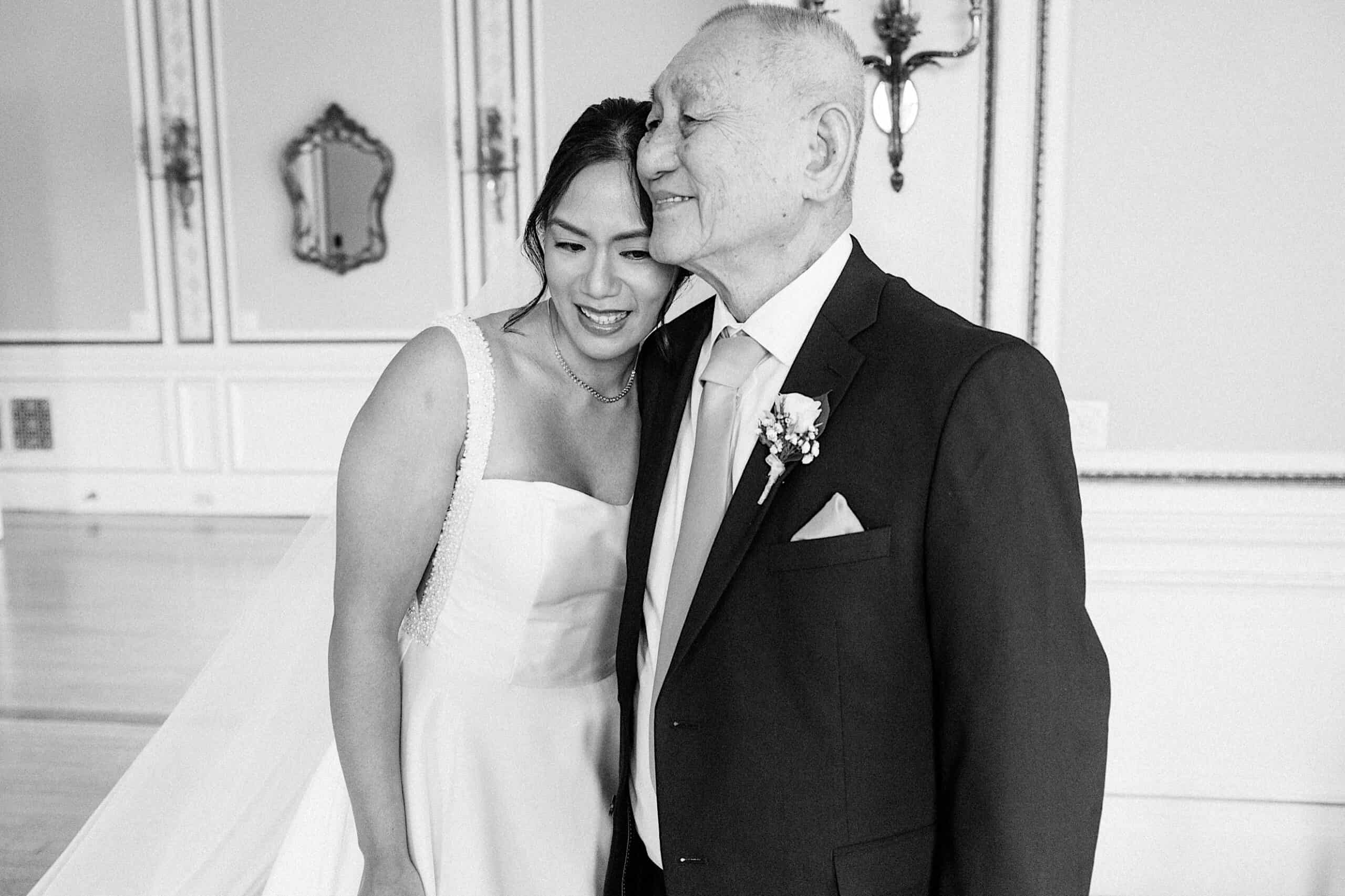 A bride in a wedding dress stands closely beside an older man in a suit, both smiling, in an elegant room with mirrors and ornate wall decor at an elegant Cairnwood Estate Wedding.