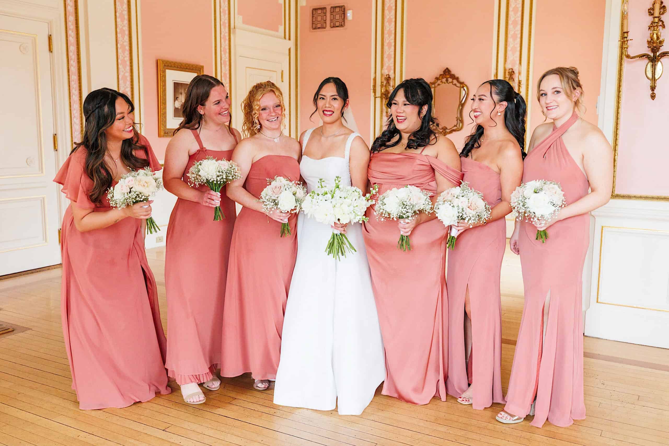 A bride in a white dress stands with six bridesmaids in matching pink dresses, all holding white bouquets, inside an elegant room with pink walls and gold accents at an elegant Cairnwood Estate wedding.