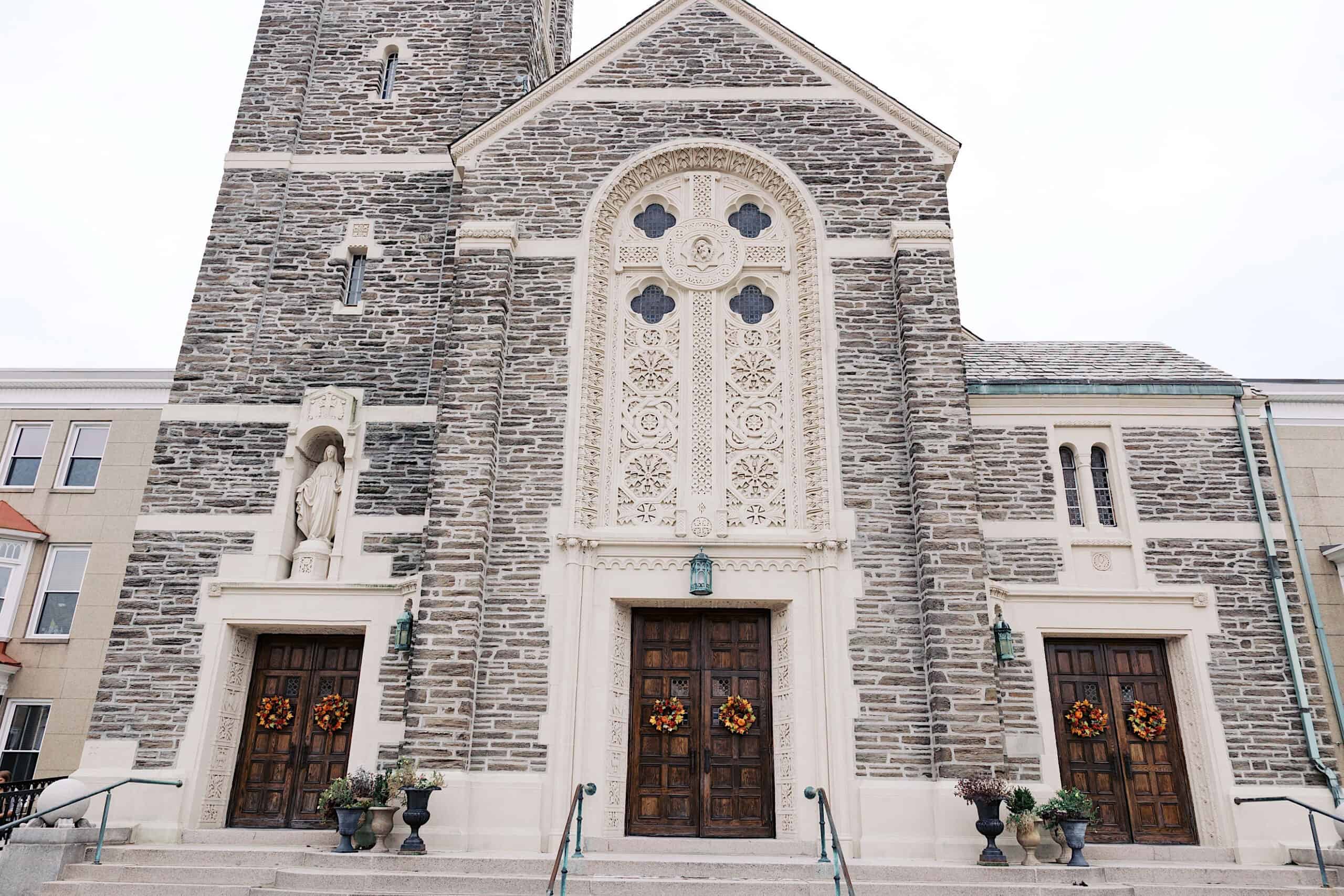 Stone church facade with three wooden doors adorned with autumn wreaths, arched windows, ornate carvings, and steps leading up to the entrances—perfect for an elegant Cairnwood Estate Wedding backdrop.