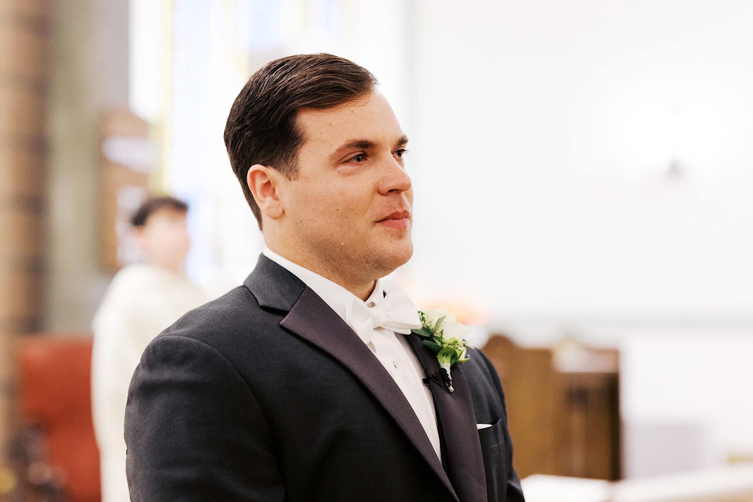 A man in a dark suit with a white bow tie and boutonniere stands indoors, looking ahead with a neutral expression at this elegant Cairnwood Estate Wedding. The background is softly lit and out of focus.