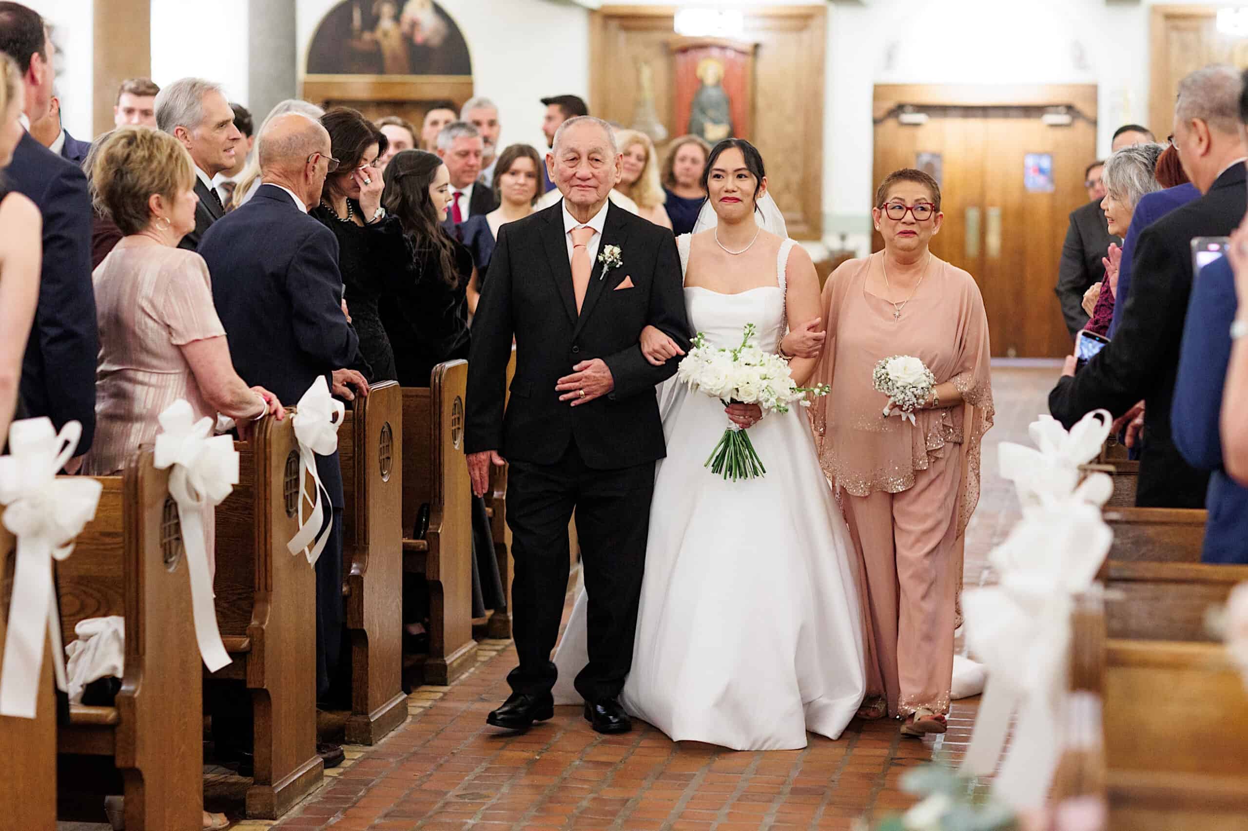 A bride in a white dress walks down the aisle with two older adults, likely her parents, as guests watch from pews at an elegant Cairnwood Estate Wedding.