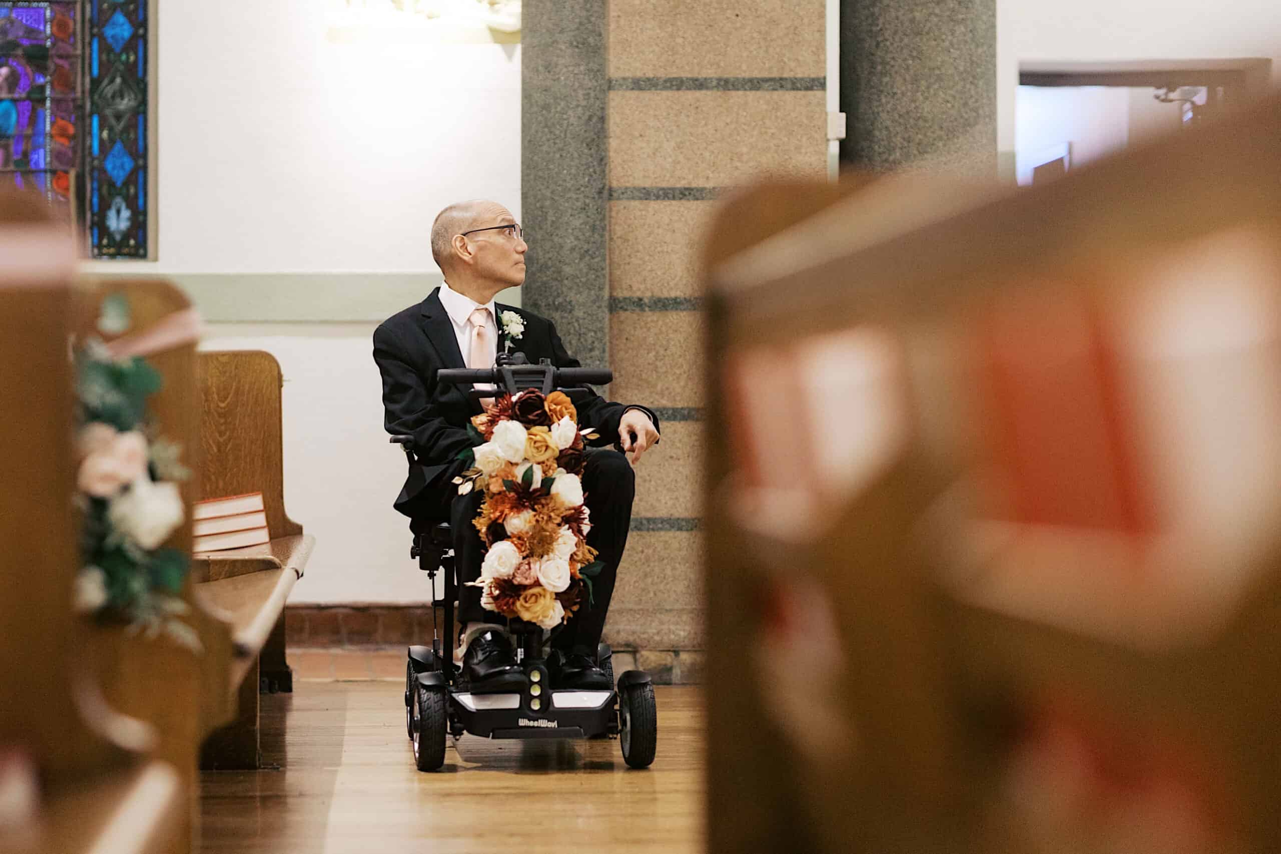 A man in a suit sits in a decorated mobility scooter inside a church, looking to the side—capturing an elegant Cairnwood Estate Wedding moment with charm and style.