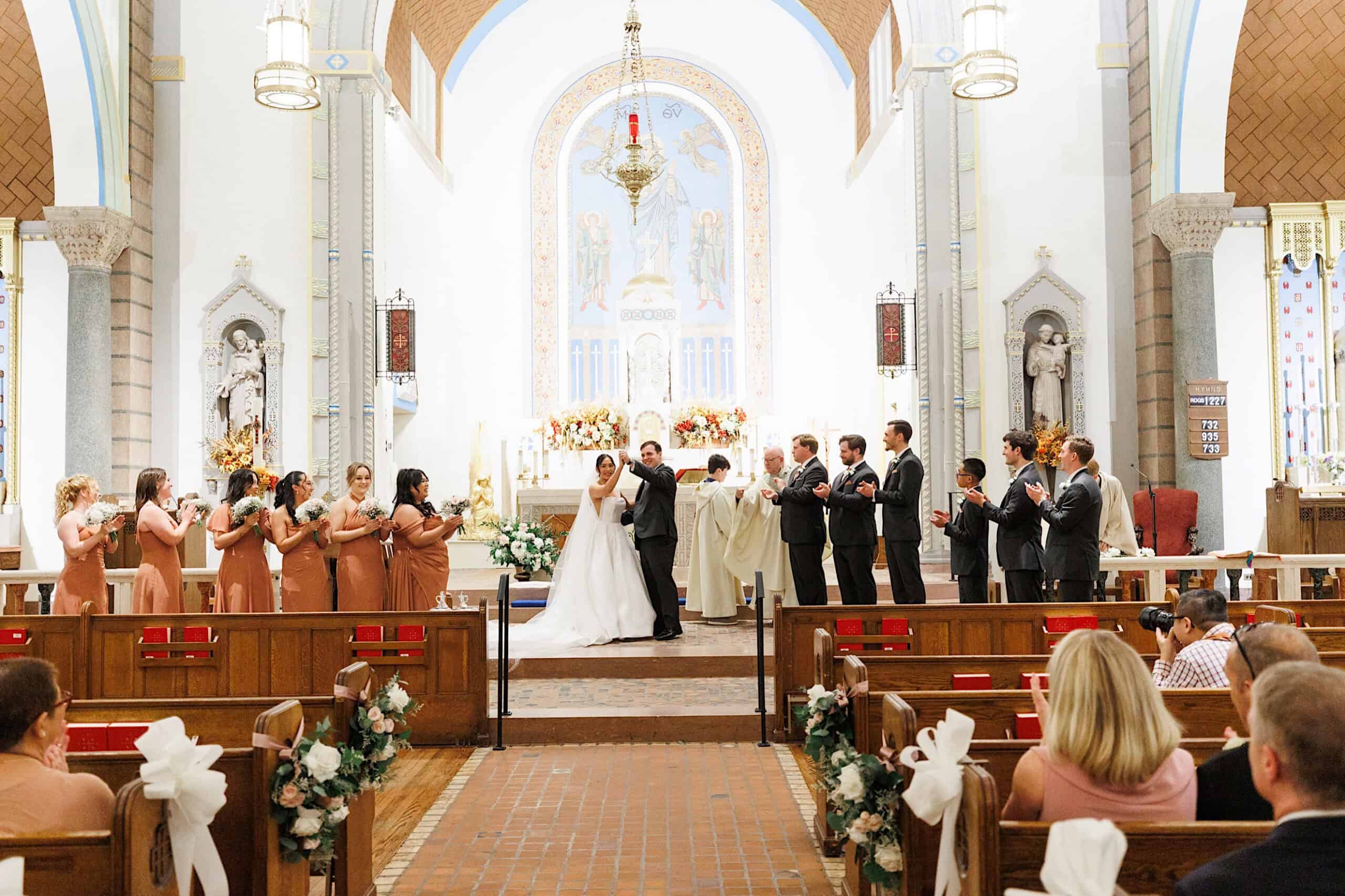 A bride and groom kiss at the altar during their elegant Cairnwood Estate Wedding ceremony, surrounded by their bridal party and guests seated in pews.