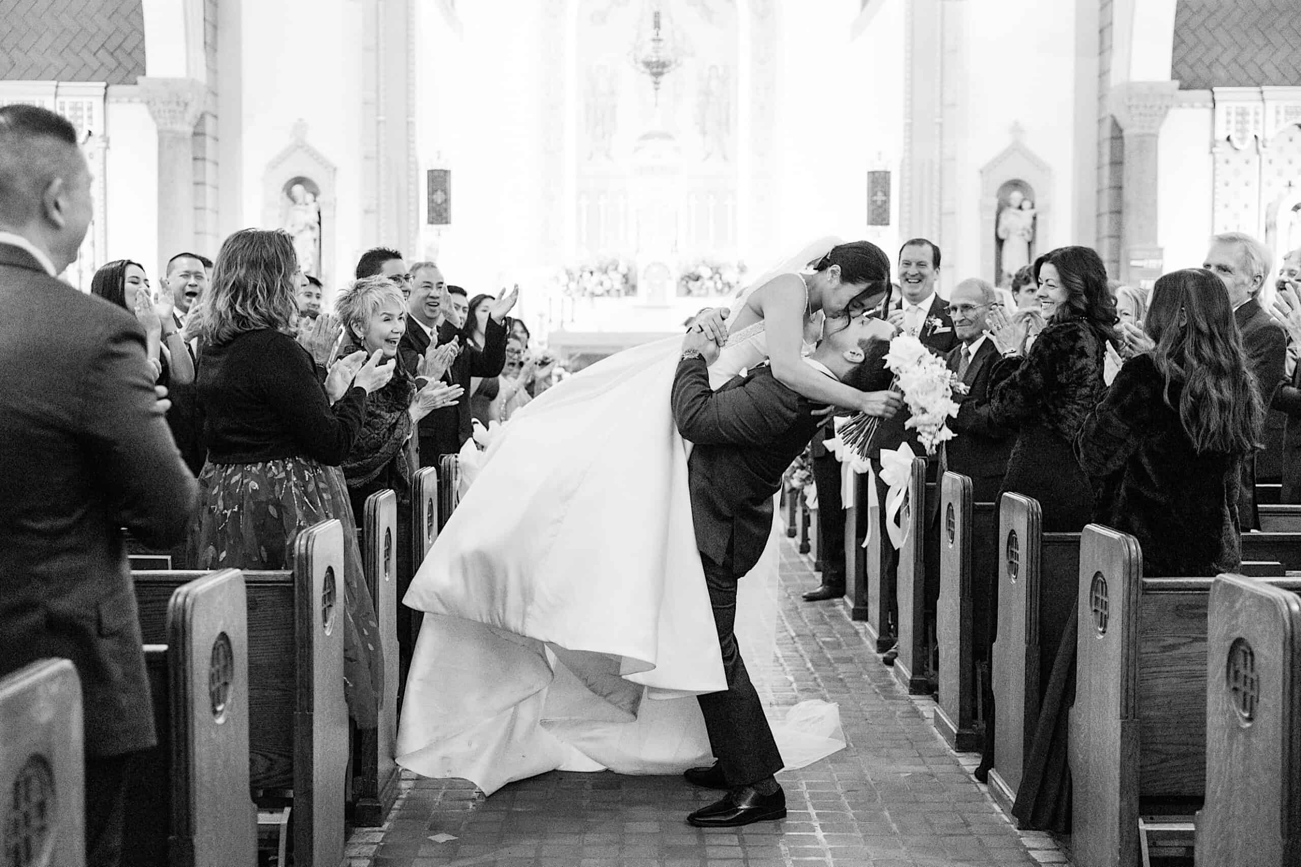 A bride and groom kiss at the end of the aisle in a church, while wedding guests standing in pews applaud and smile, evoking the romantic charm of an elegant Cairnwood Estate Wedding.