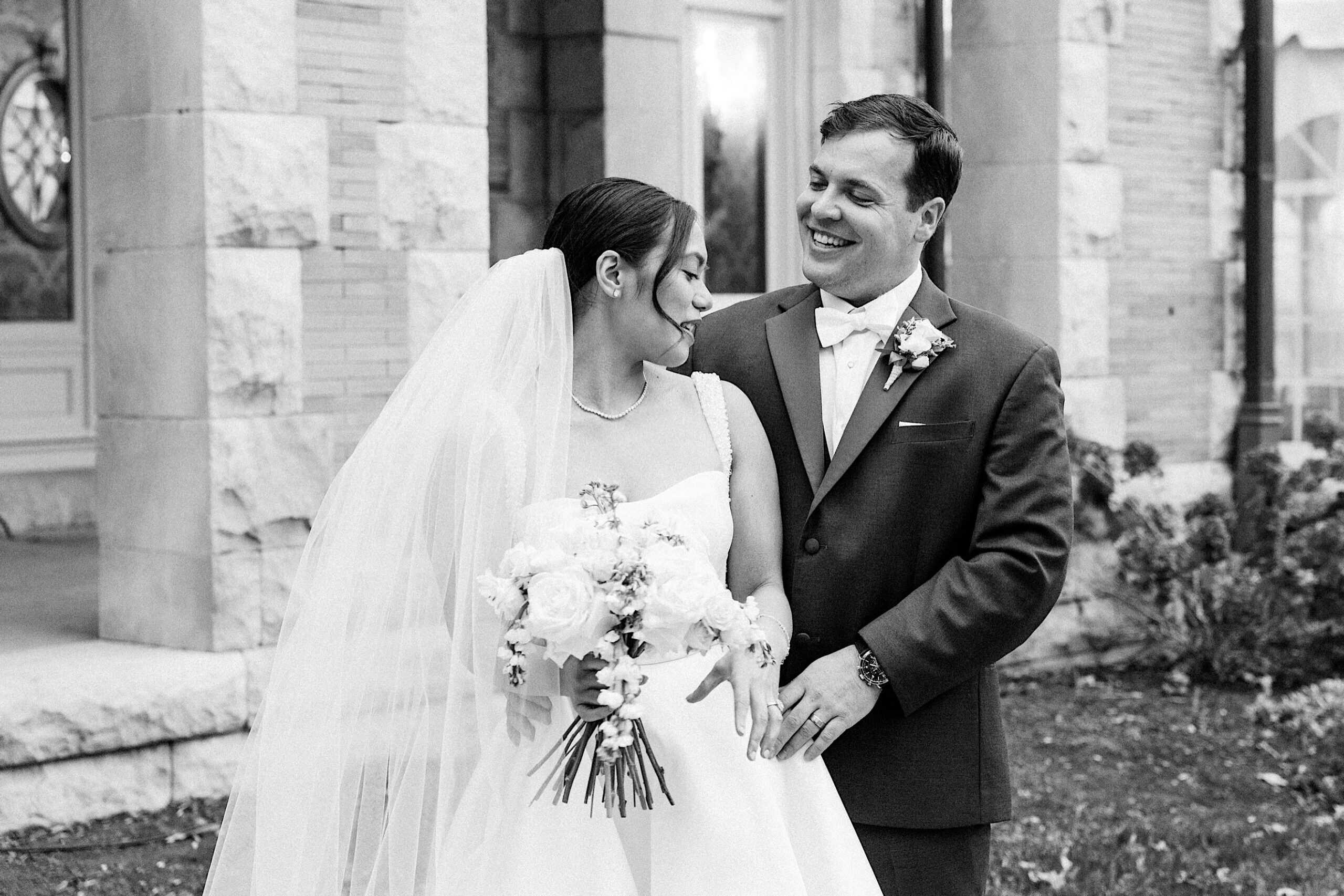 A bride and groom stand together outside a stone building at their elegant Cairnwood Estate wedding, smiling at each other. The bride holds a bouquet and wears a veil, while the groom wears a suit with a bow tie.