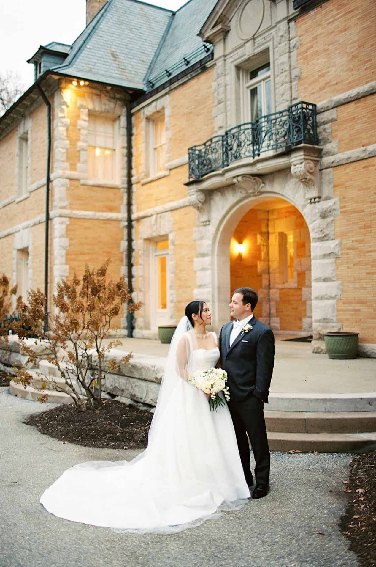 A bride and groom pose together outside the elegant Cairnwood Estate, both smiling; the bride holds a bouquet of white flowers.