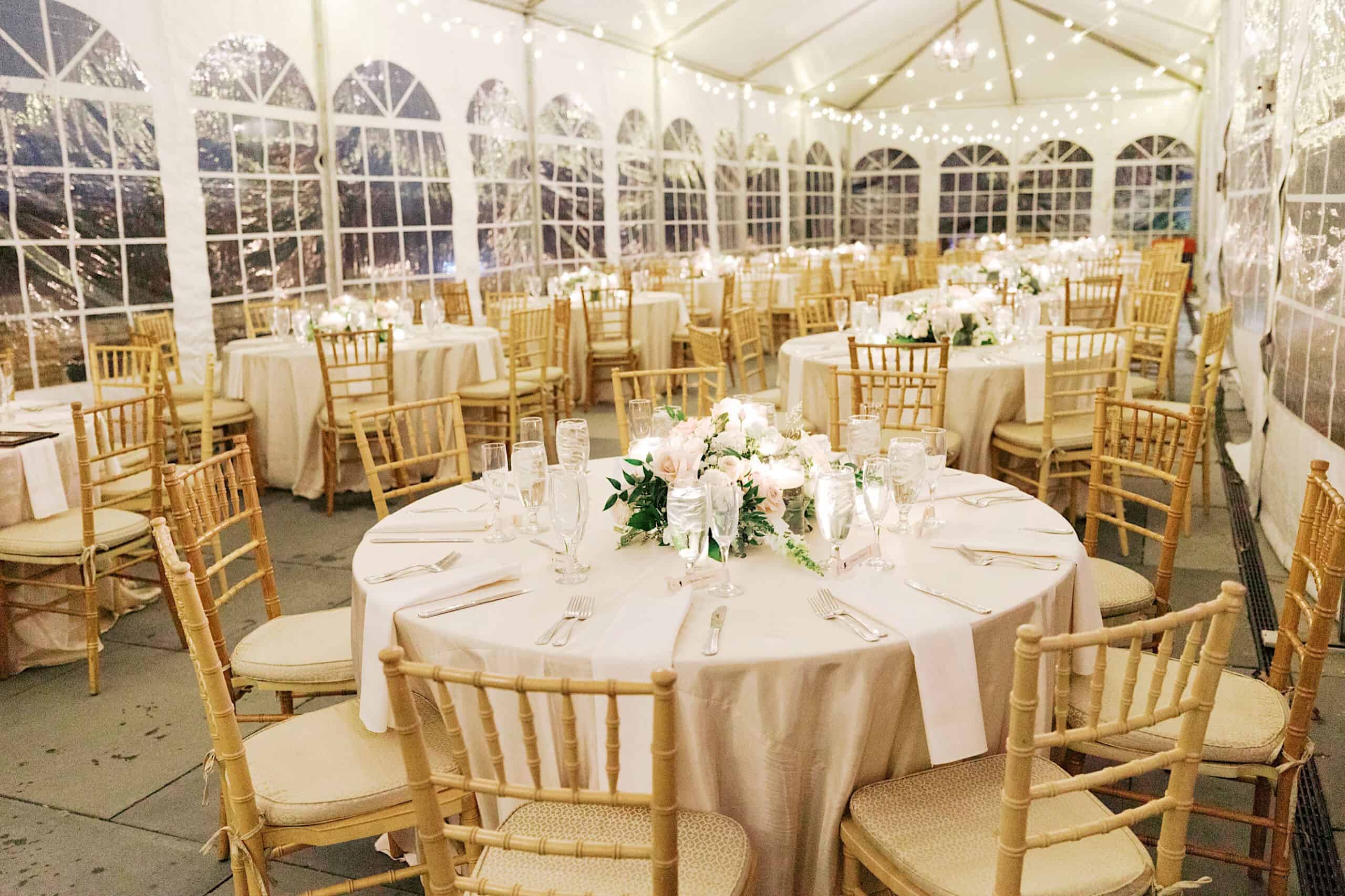 Elegant Cairnwood Estate Wedding reception setup under a tent with round tables, beige tablecloths, floral centerpieces, and wooden chairs, all beautifully illuminated by string lights.