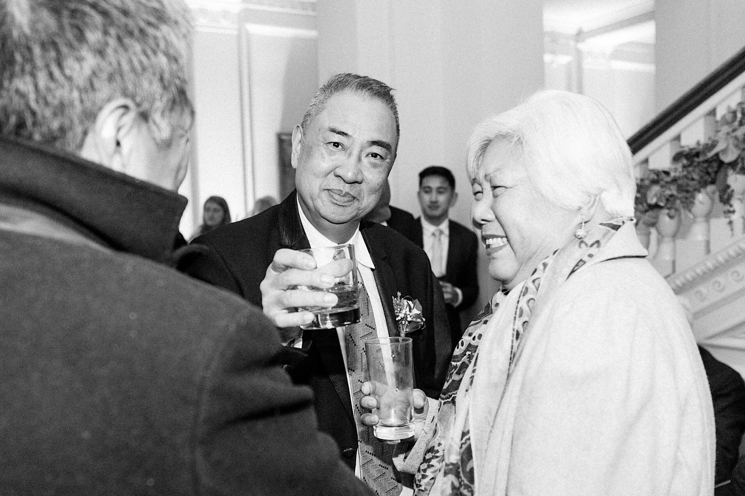 At an elegant Cairnwood Estate Wedding, a man holding a drink raises his glass toward the camera while standing next to a smiling woman at an indoor social gathering.