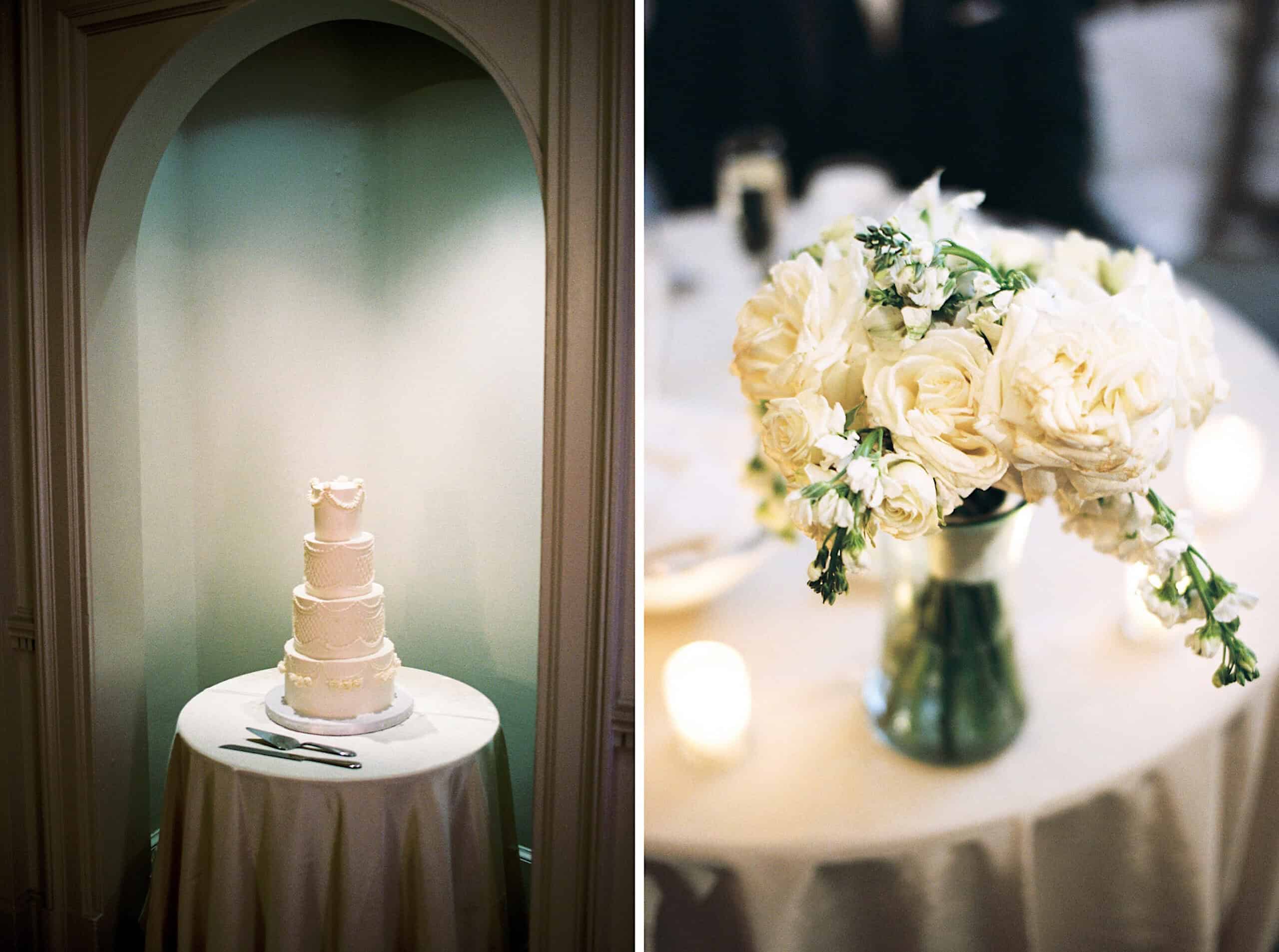 A four-tiered white wedding cake on a round table at an elegant Cairnwood Estate Wedding, set beside a silver vase of white flowers and glowing candles on another table.