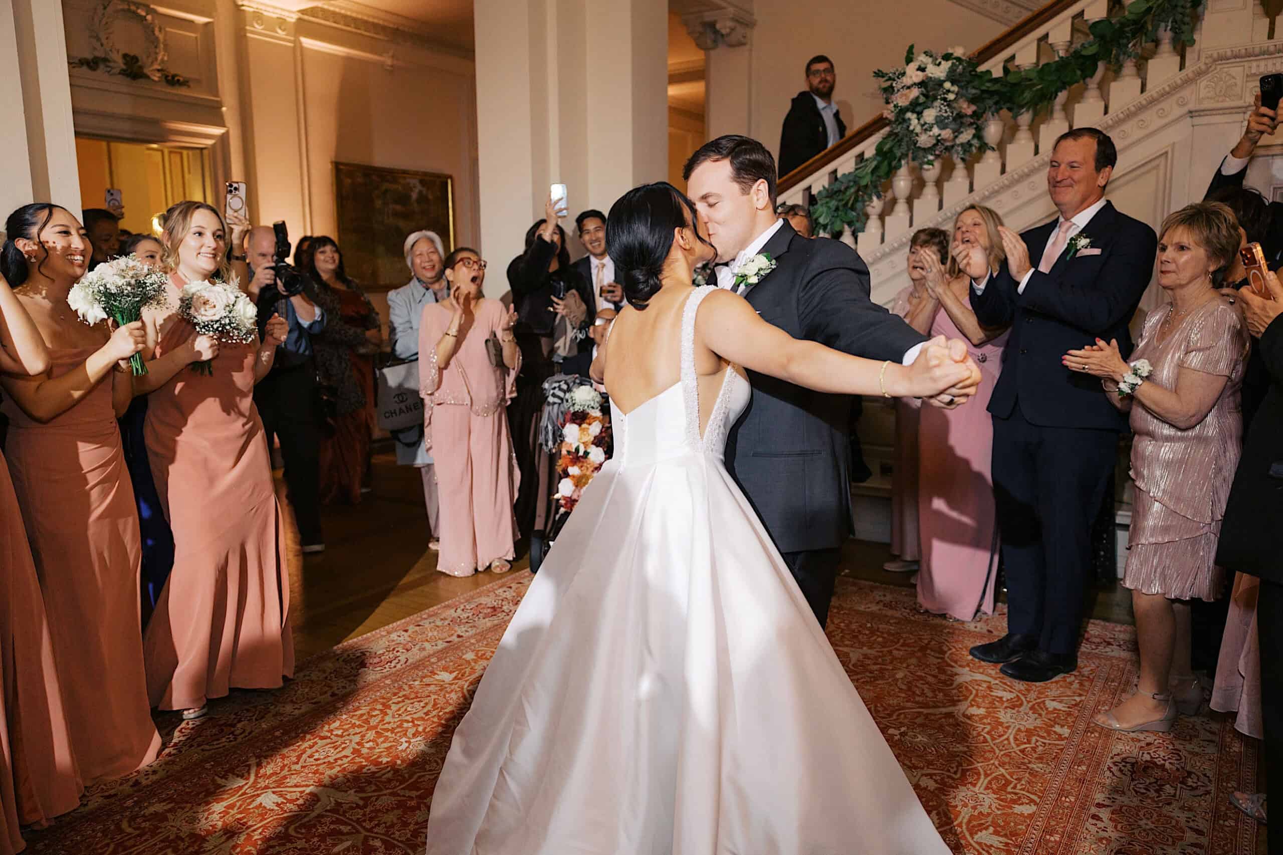 A bride and groom share their first dance in a formal indoor setting at an elegant Cairnwood Estate Wedding, surrounded by guests in formal attire, some holding bouquets and clapping.