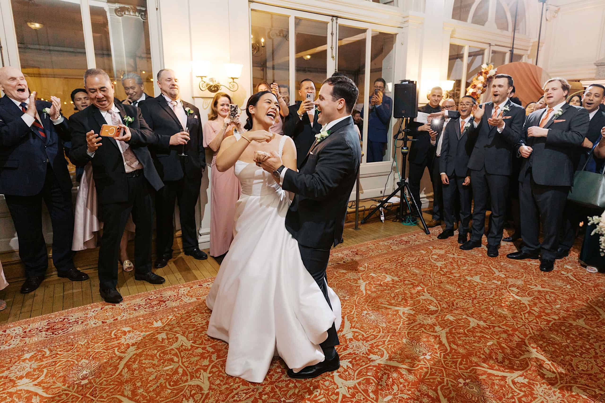 A bride and groom share a joyful dance on a patterned carpet at their elegant Cairnwood Estate Wedding, smiling as guests in formal attire watch and capture the special moment.